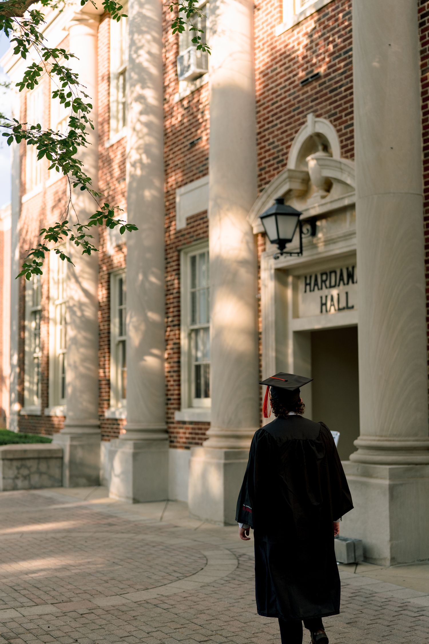 Graduate in cap and gown walking toward Hardaway Hall brick building with white columns.