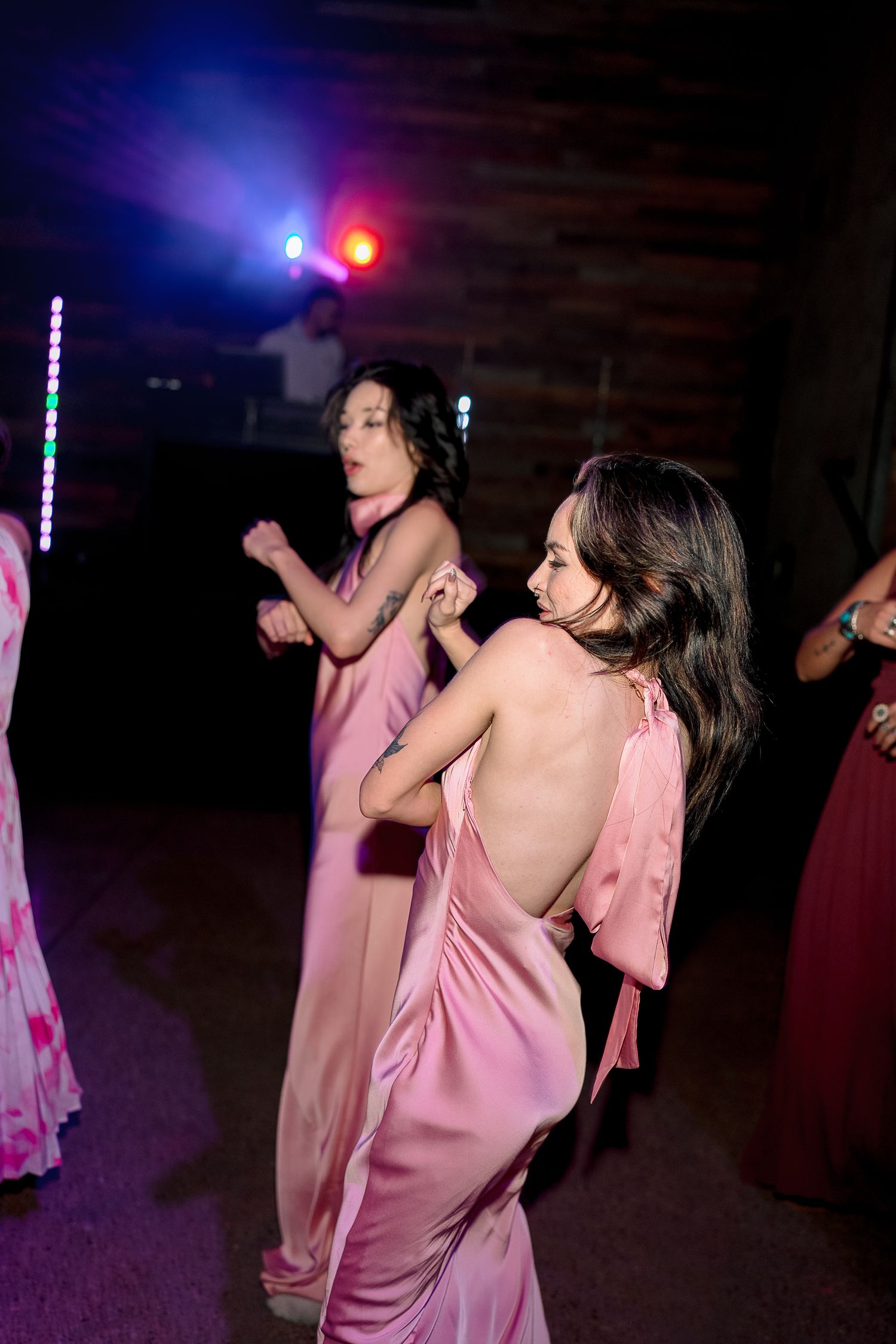 Bridesmaids in pink satin halter gowns dancing at wedding reception under colorful DJ lights.