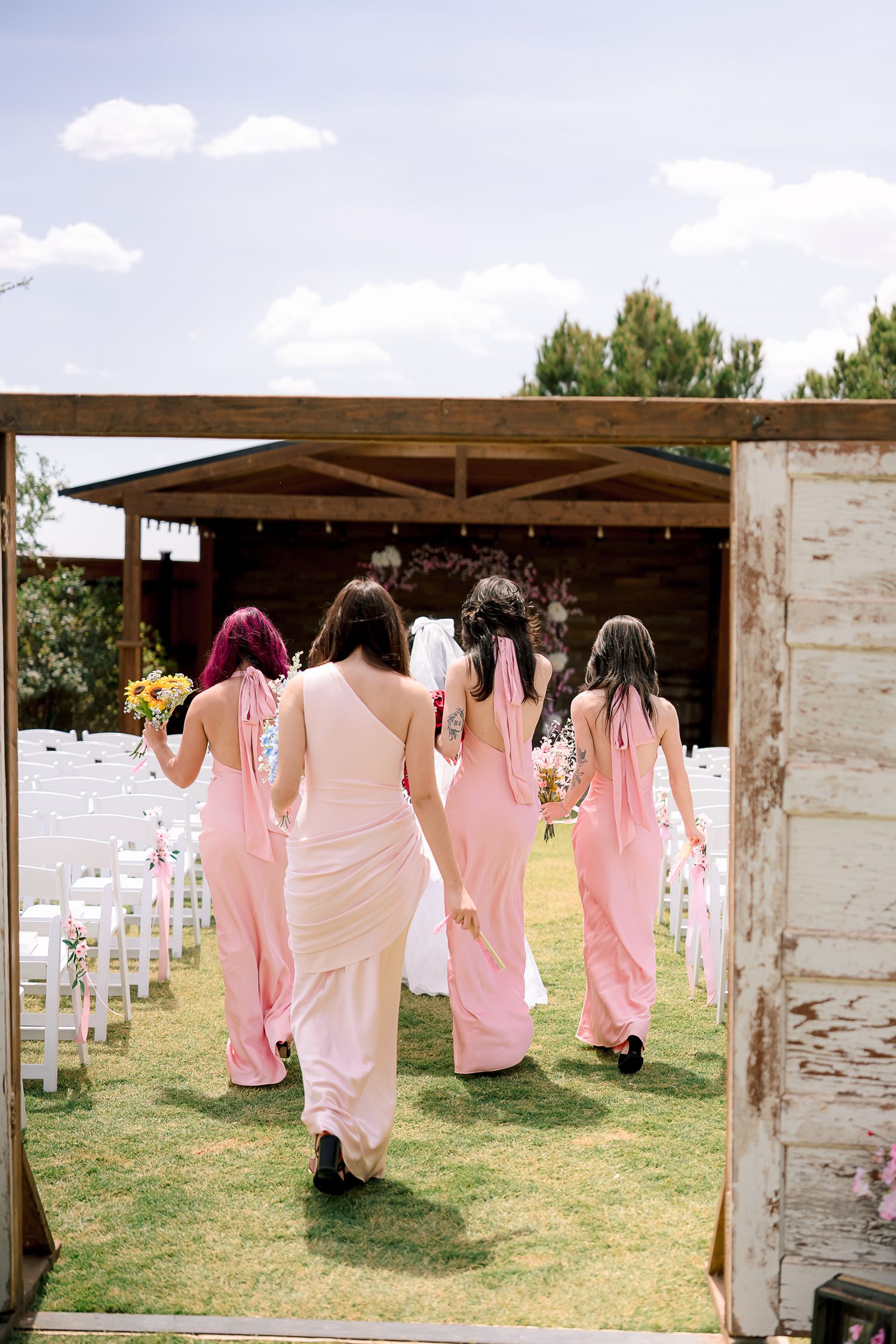 Bridesmaids in pink dresses walk down outdoor wedding aisle toward rustic wooden ceremony venue.