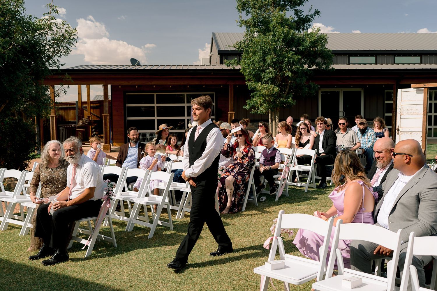 Wedding guests seated in white chairs watch groomsman walk down outdoor ceremony aisle.