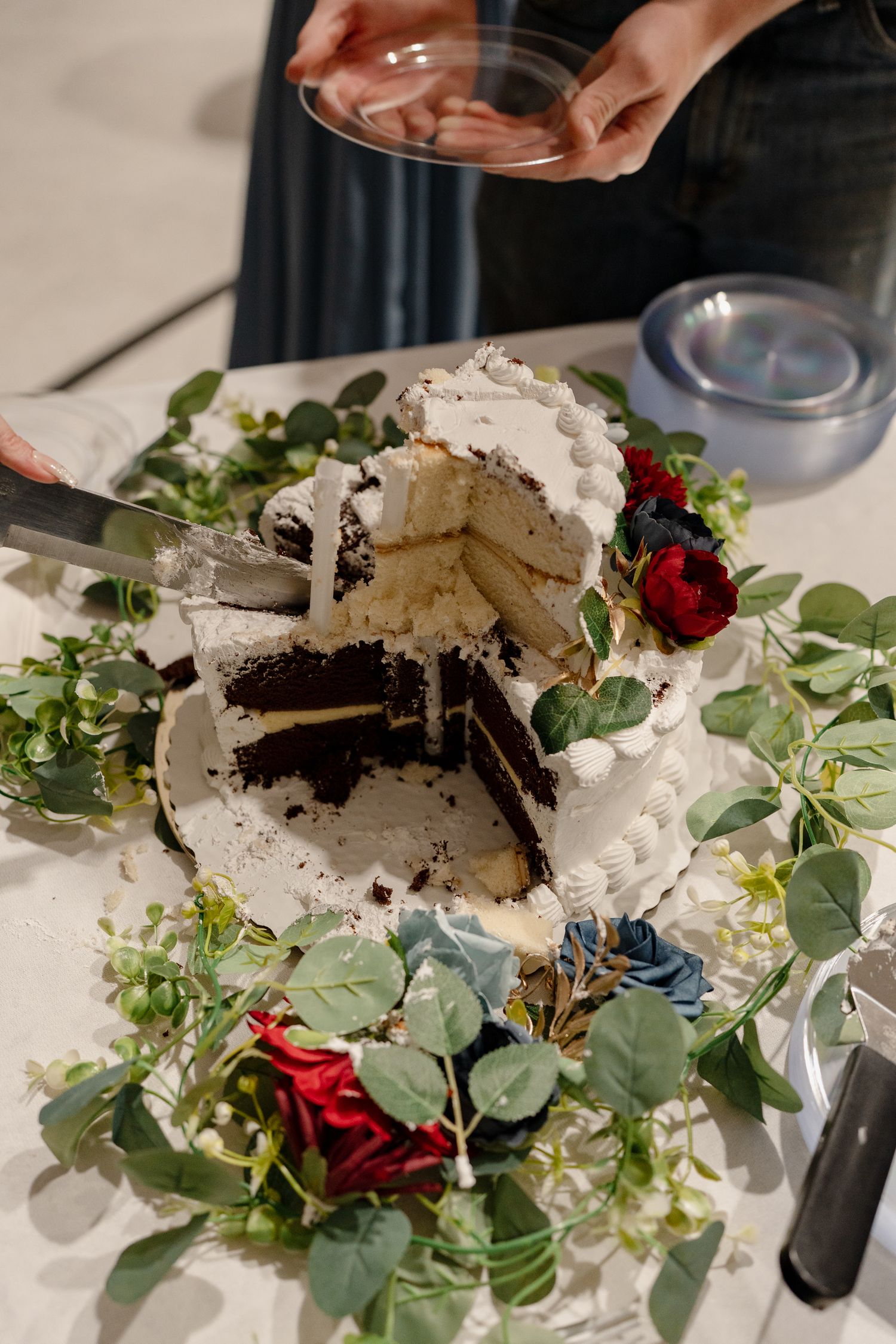 A chocolate cake with white frosting being cut and served on a table decorated with eucalyptus leaves and red roses.