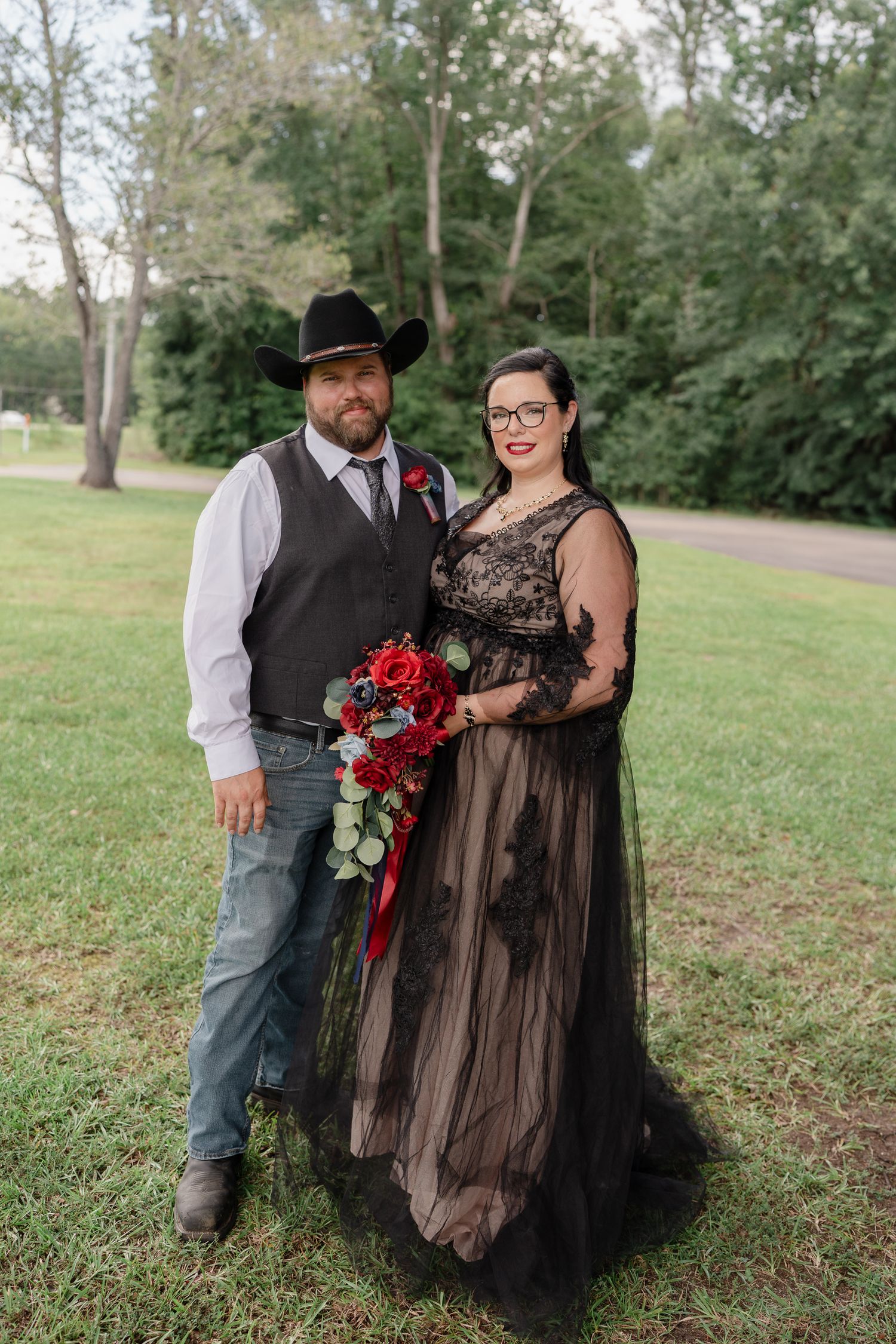 A couple poses in a grassy field wearing a black tulle gown and western attire with red roses.
