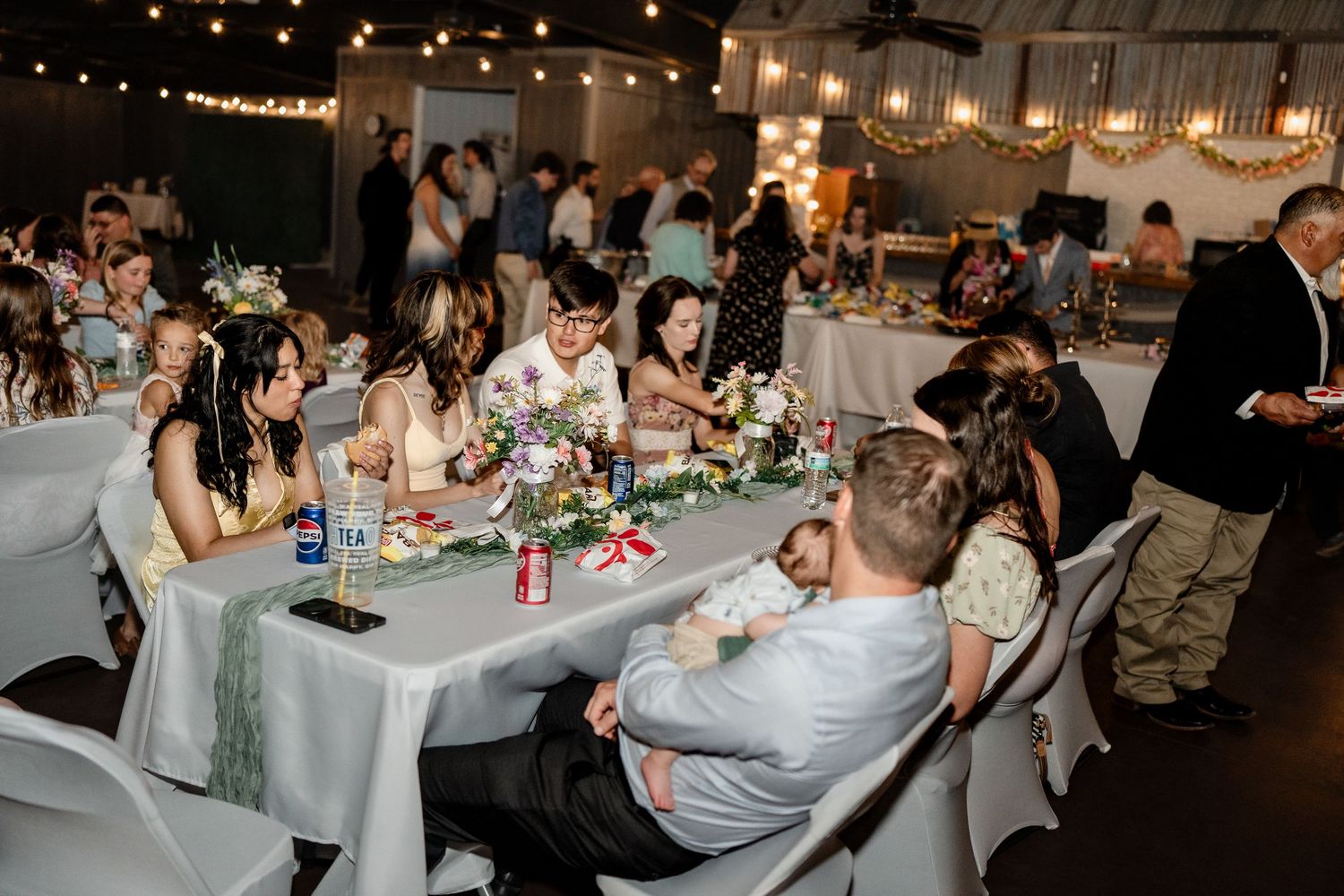 Wedding reception guests seated at long tables in a dimly lit venue with string lights and festive decorations.