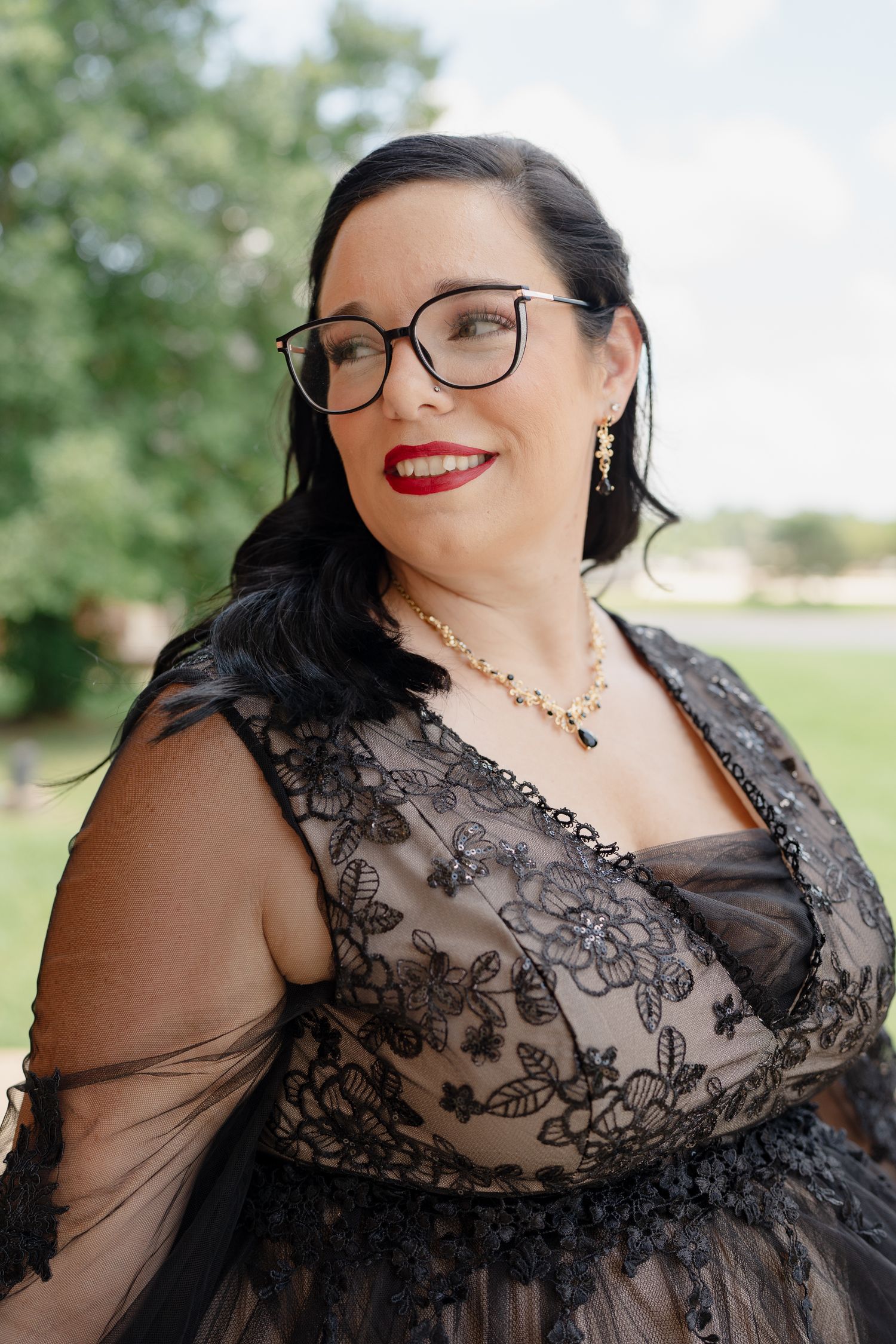 A woman in a black lace formal dress with jewelry and glasses poses outdoors.
