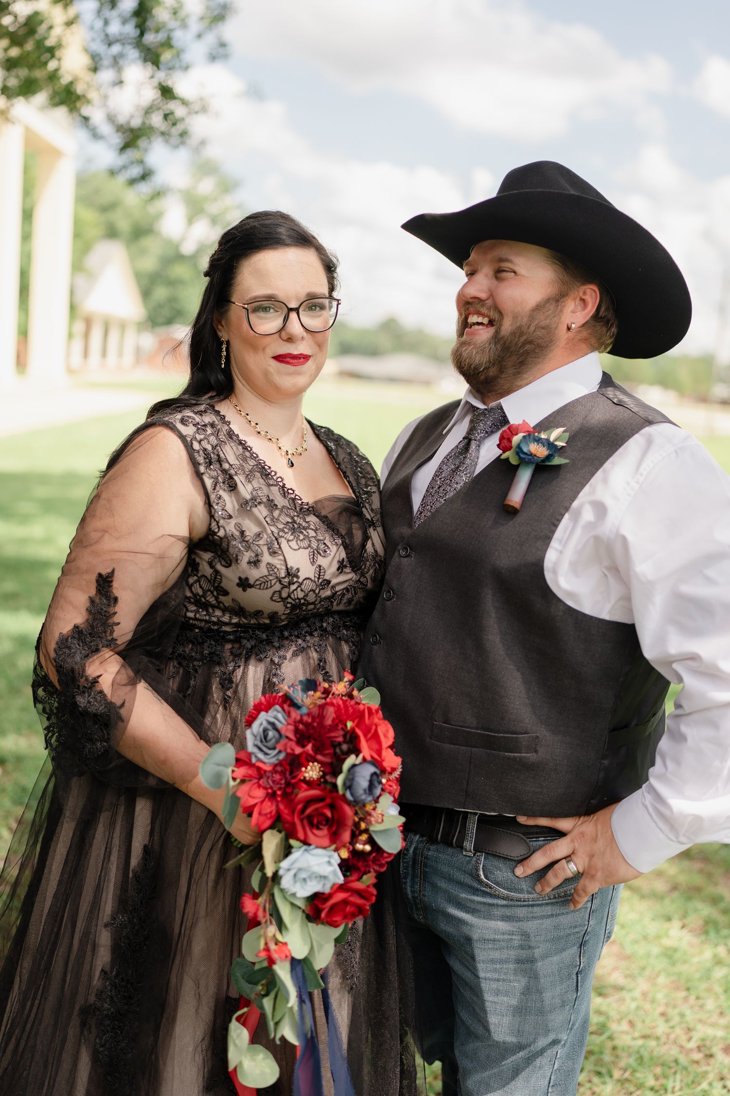 A couple in black western-themed wedding attire poses with a vibrant red bouquet outdoors.