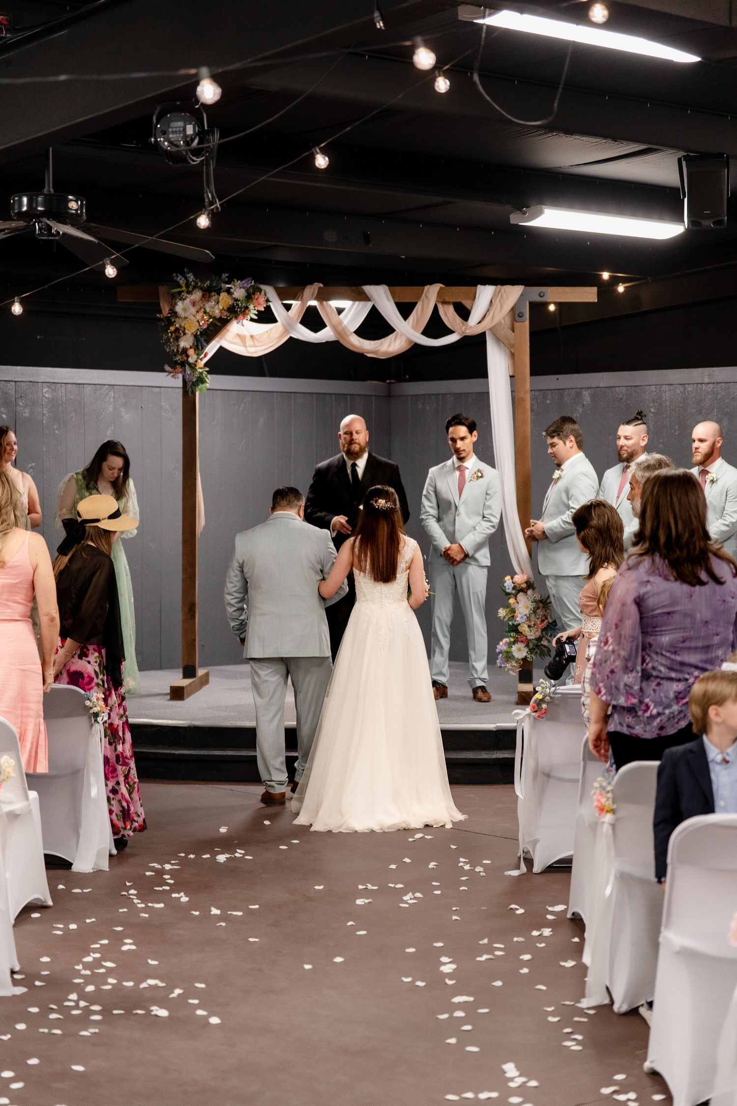 Wedding ceremony in progress with guests gathered in an industrial-style venue decorated with white draping and rose petals.