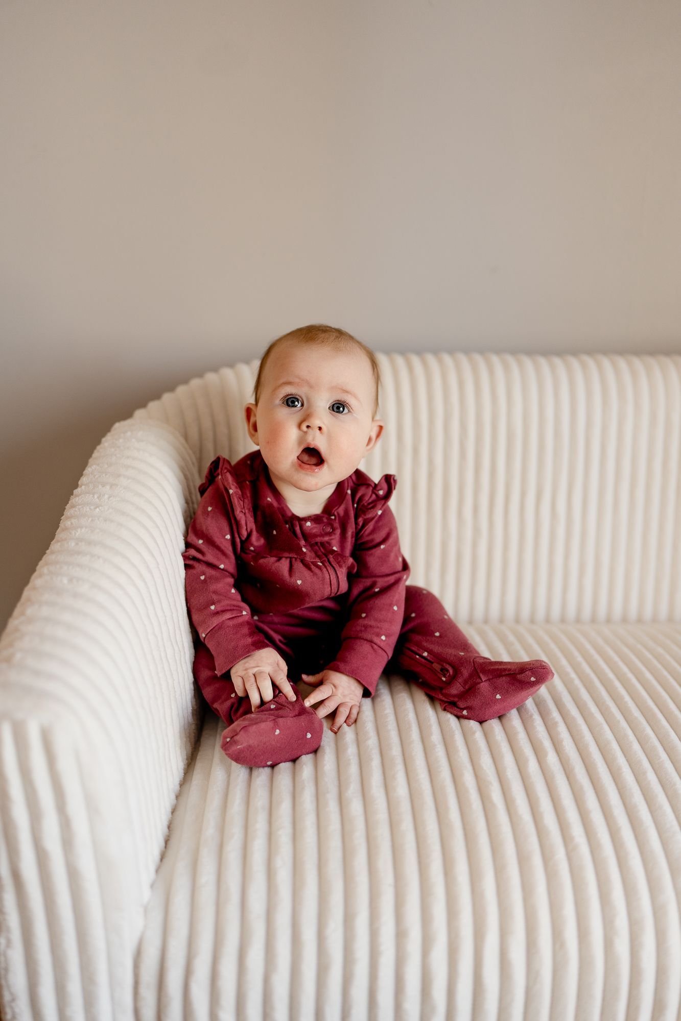 Baby in burgundy outfit crawling on white textured sofa.