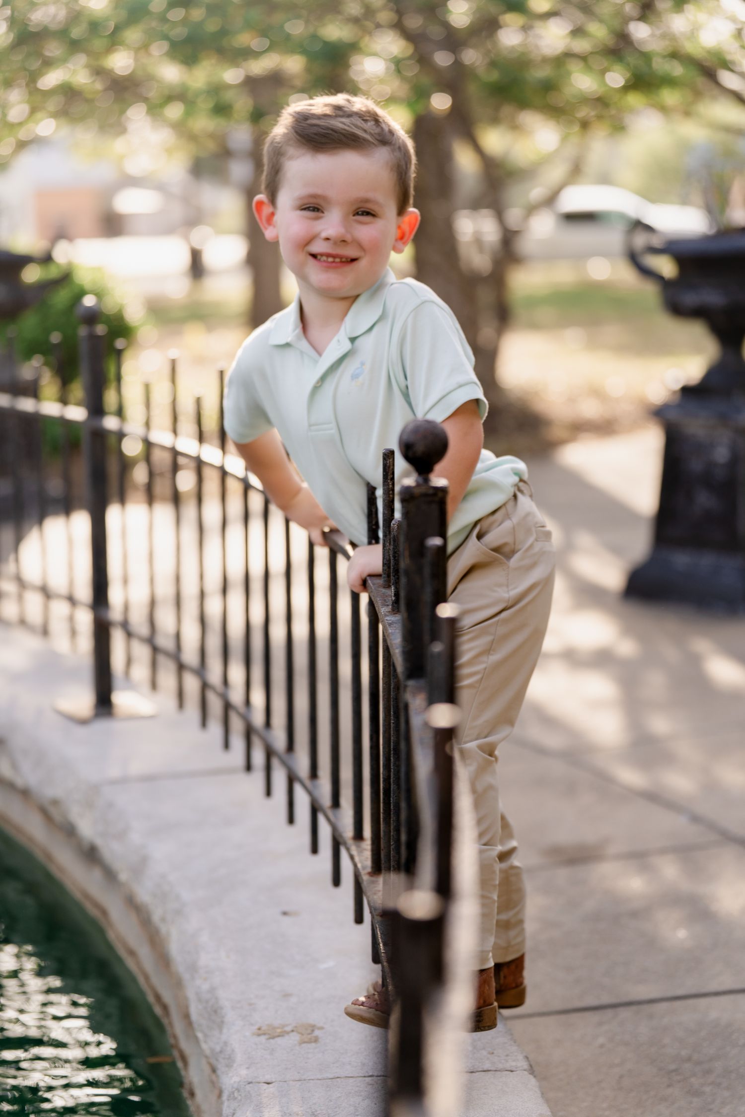 A child in casual attire poses playfully against an iron fence near water.
