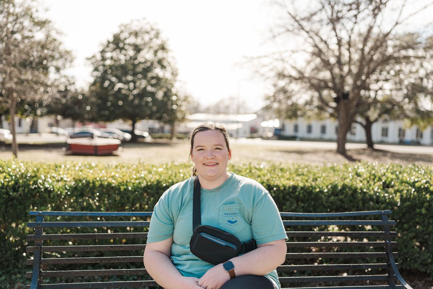 Person in teal shirt sits on park bench with trees and buildings in background.