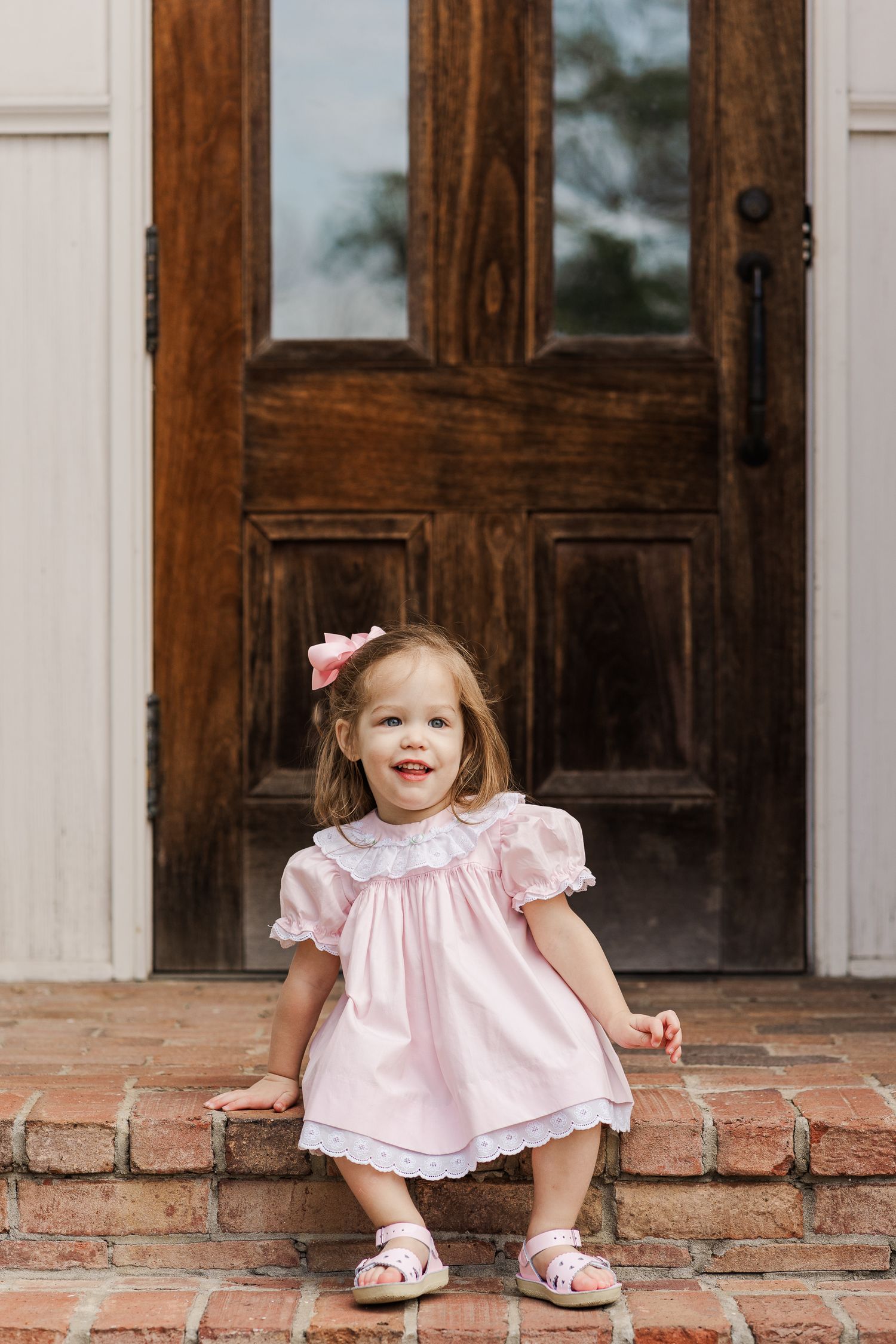 Child in pink ruffled dress crouches playfully on brick steps near wooden door.