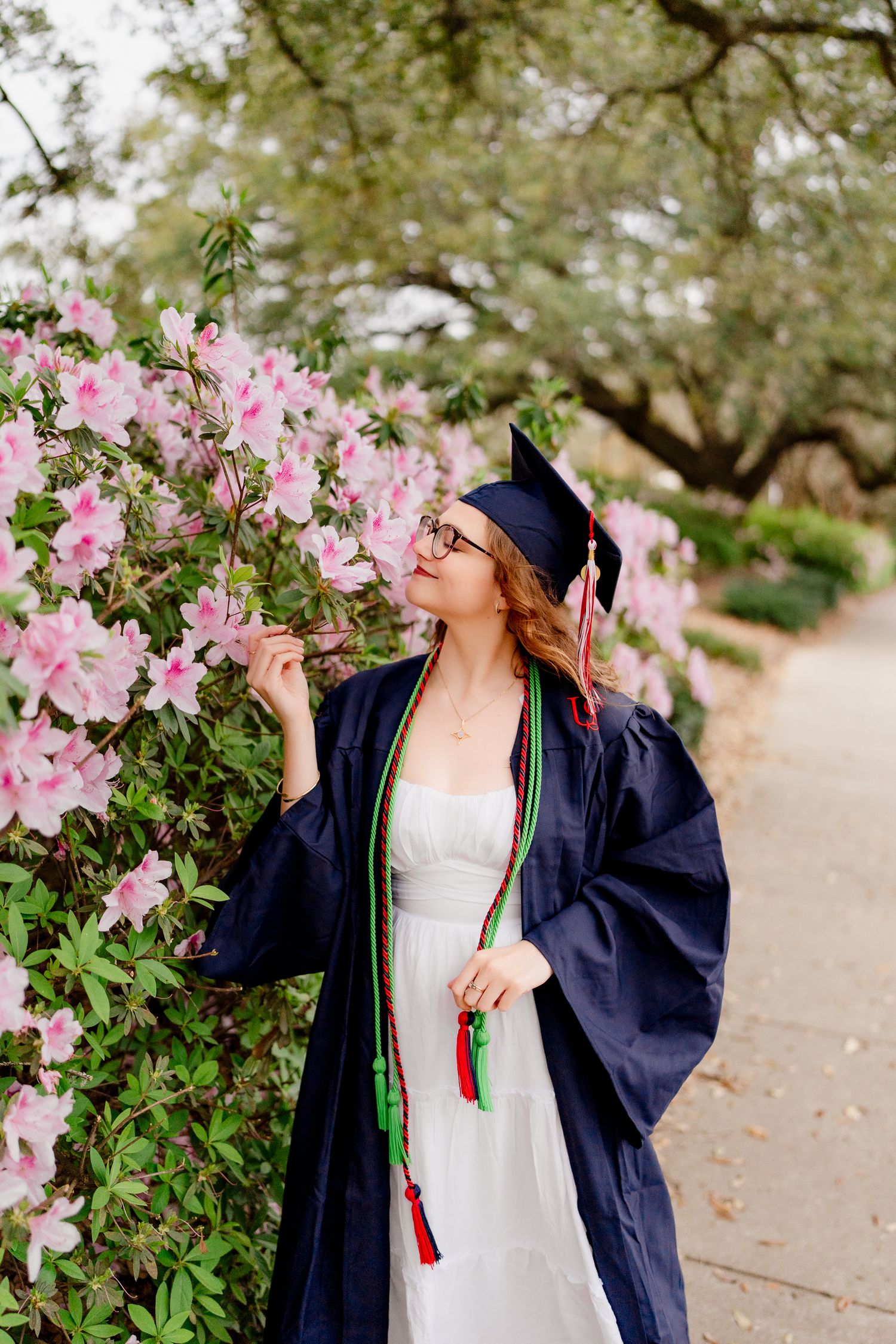 Graduate in cap and gown admires blooming pink azalea flowers on campus.