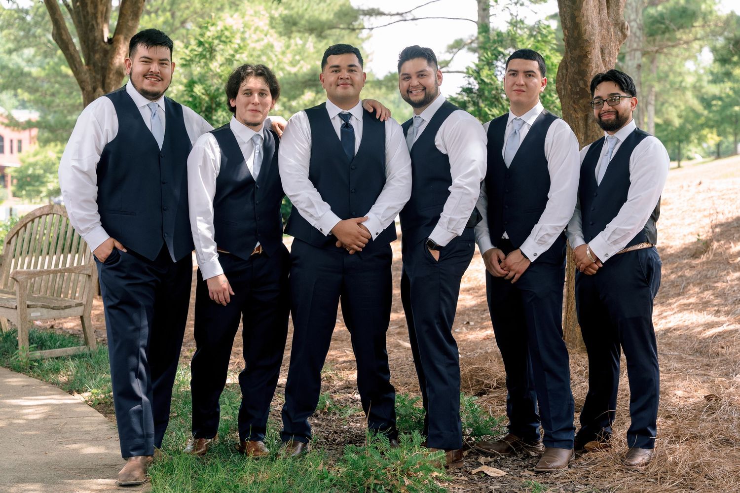 A wedding party in matching navy vests and white shirts poses together outdoors in a garden setting.