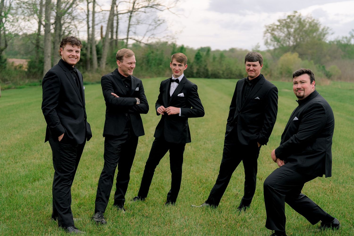 Five young men in black suits posing outdoors on green grass for a formal group photo.