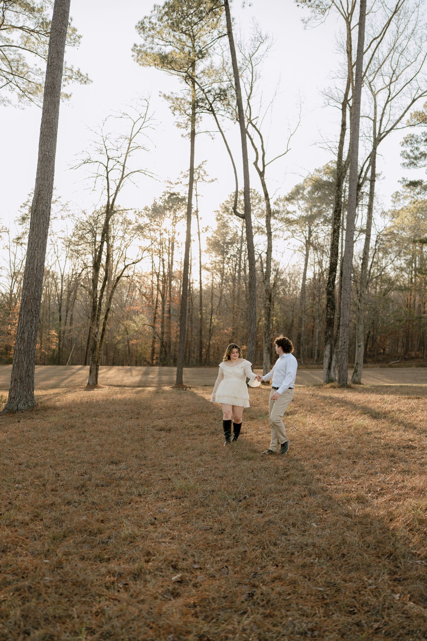 Couple dancing playfully in a sunlit pine forest clearing, woman in white dress and black boots spinning joyfully.