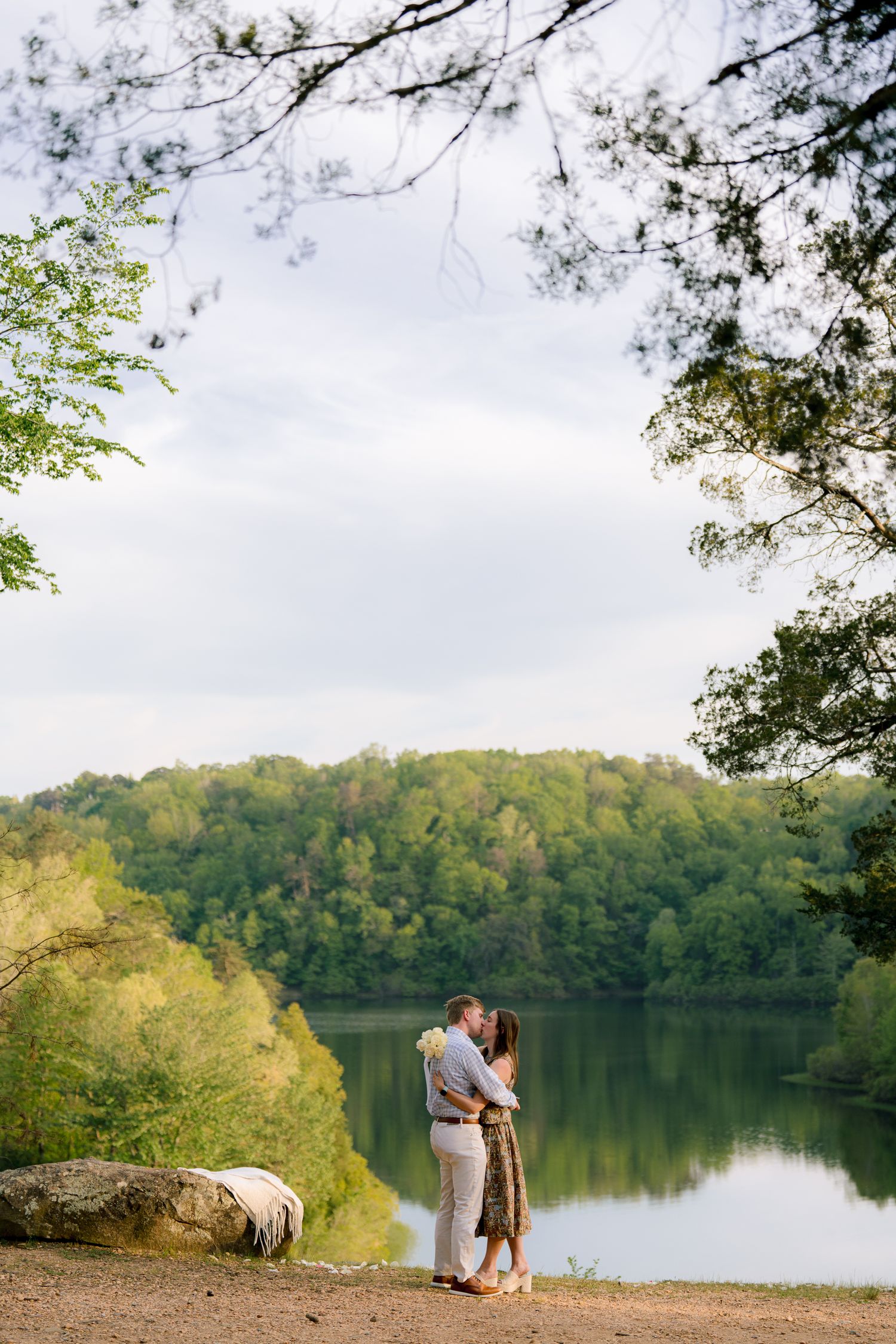 Couple kissing by a serene lake surrounded by lush green forest trees in a romantic outdoor engagement photo.