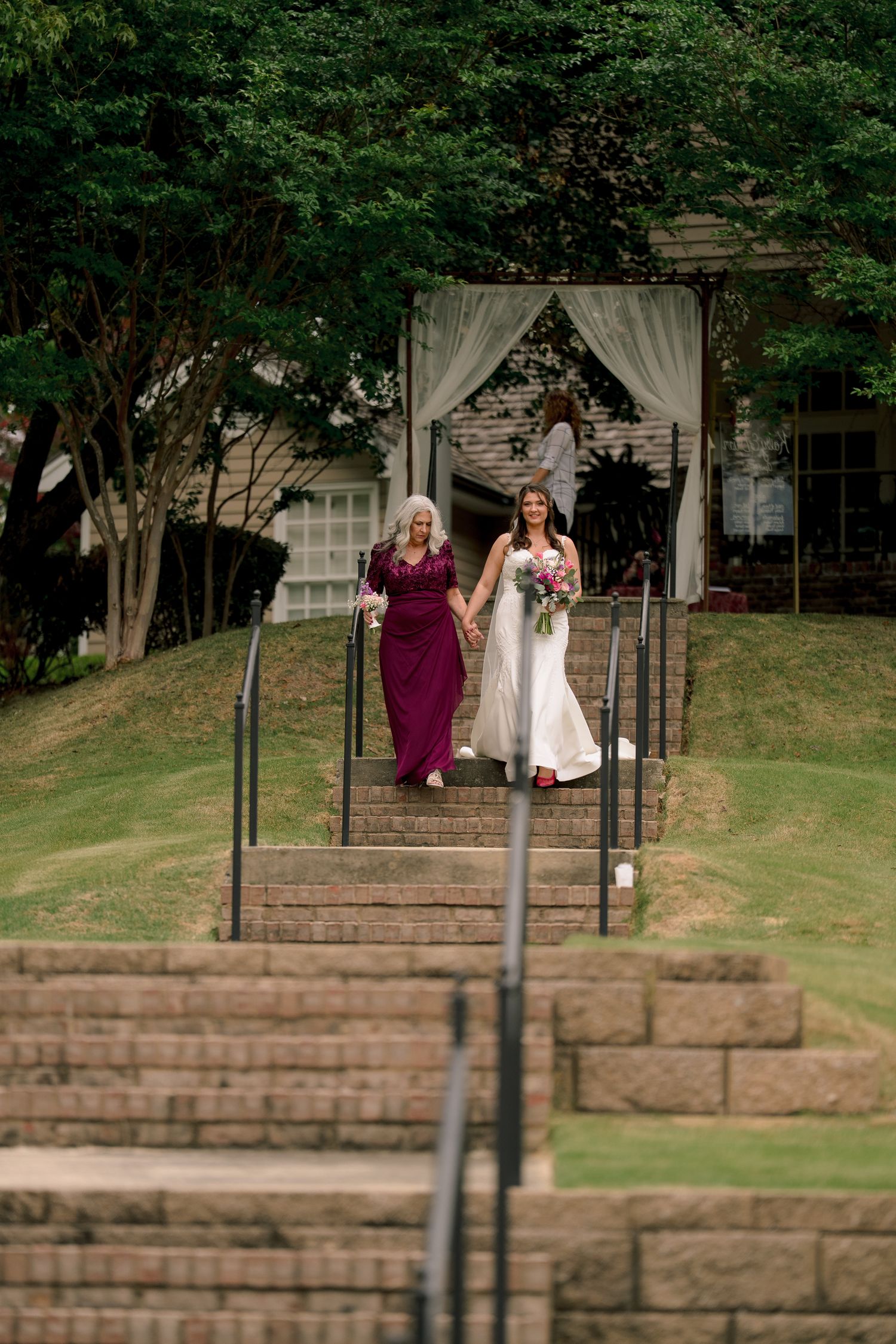Bride in white gown walks down outdoor steps with bridesmaid in burgundy dress at a garden wedding venue.