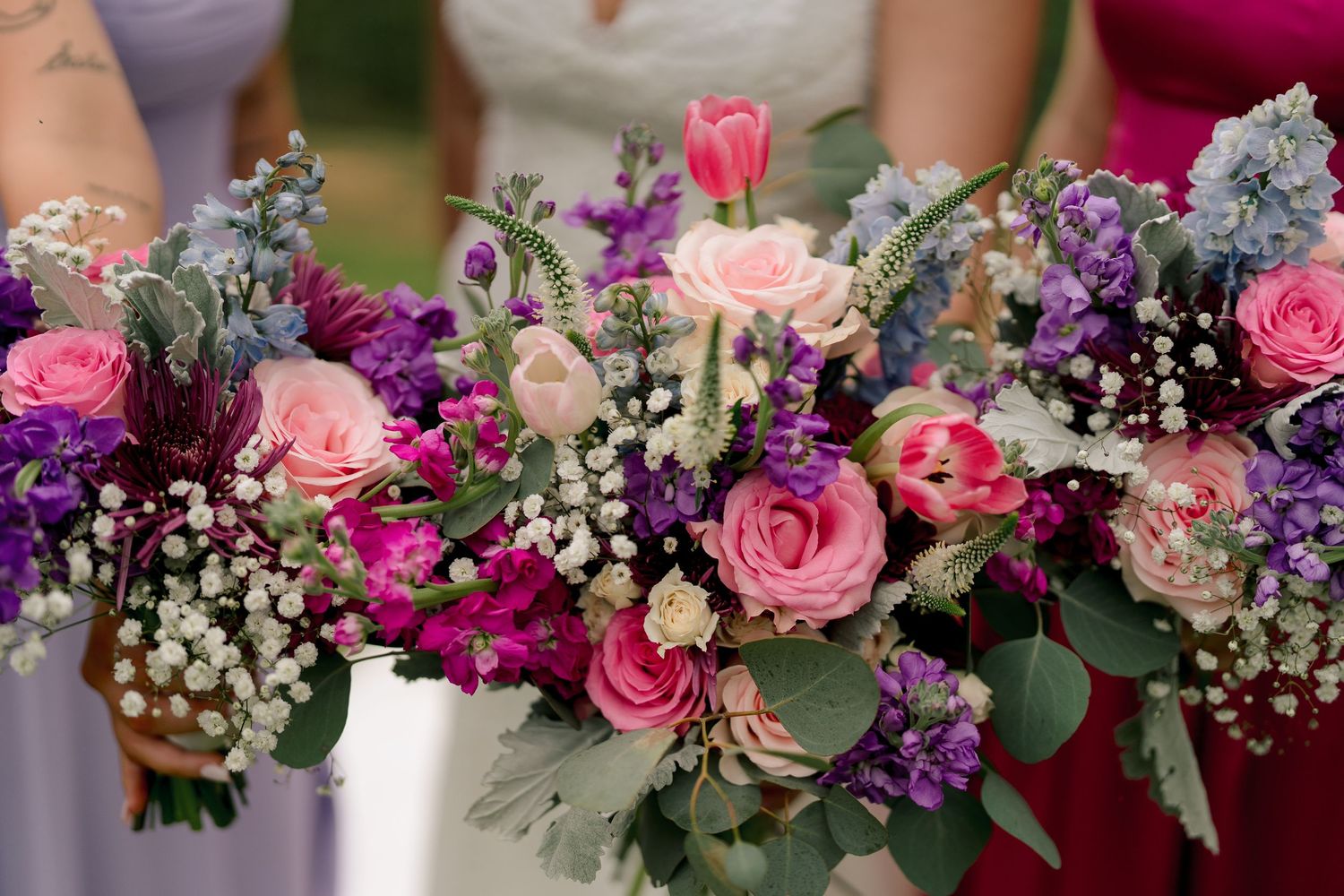 Lush bridal bouquet featuring pink roses, purple flowers, tulips, and baby's breath held by bridesmaids in colorful dresses.