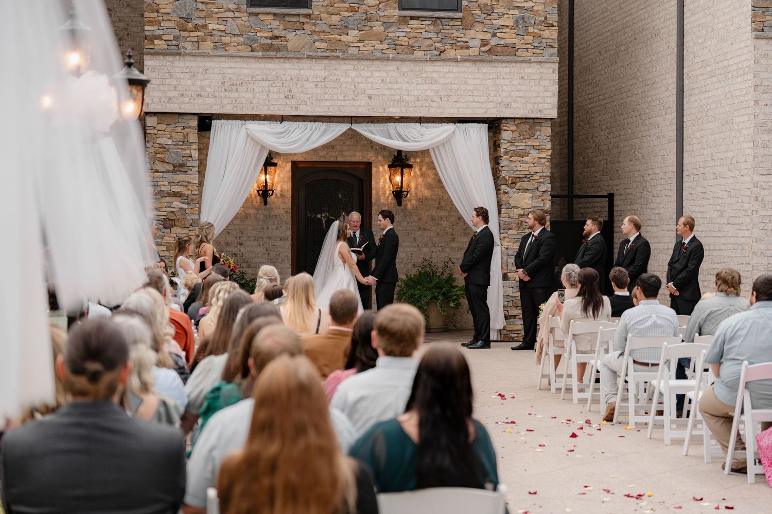 An outdoor wedding ceremony takes place at a rustic stone venue with white chairs and scattered rose petals down the aisle.