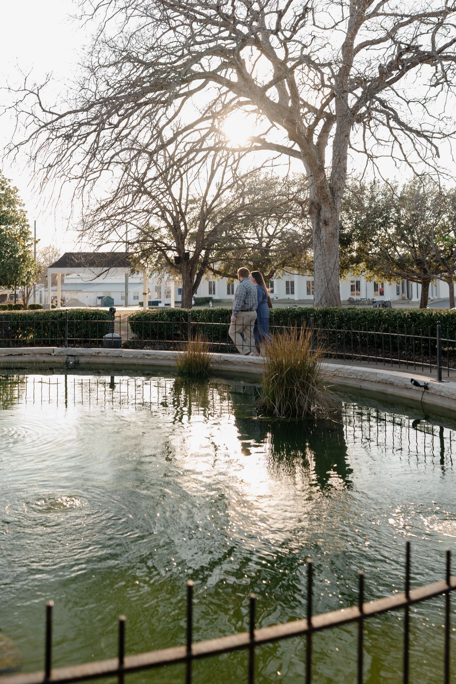 Couple standing by a reflective pond in a sunlit park with bare trees and white buildings in the background.