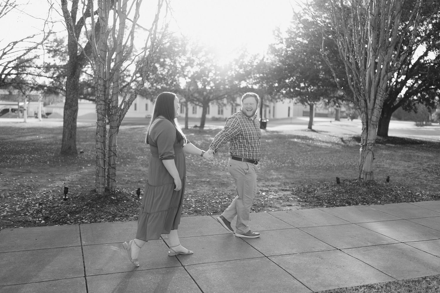 Black and white image of a couple holding hands and walking together on a sunlit sidewalk among bare trees.