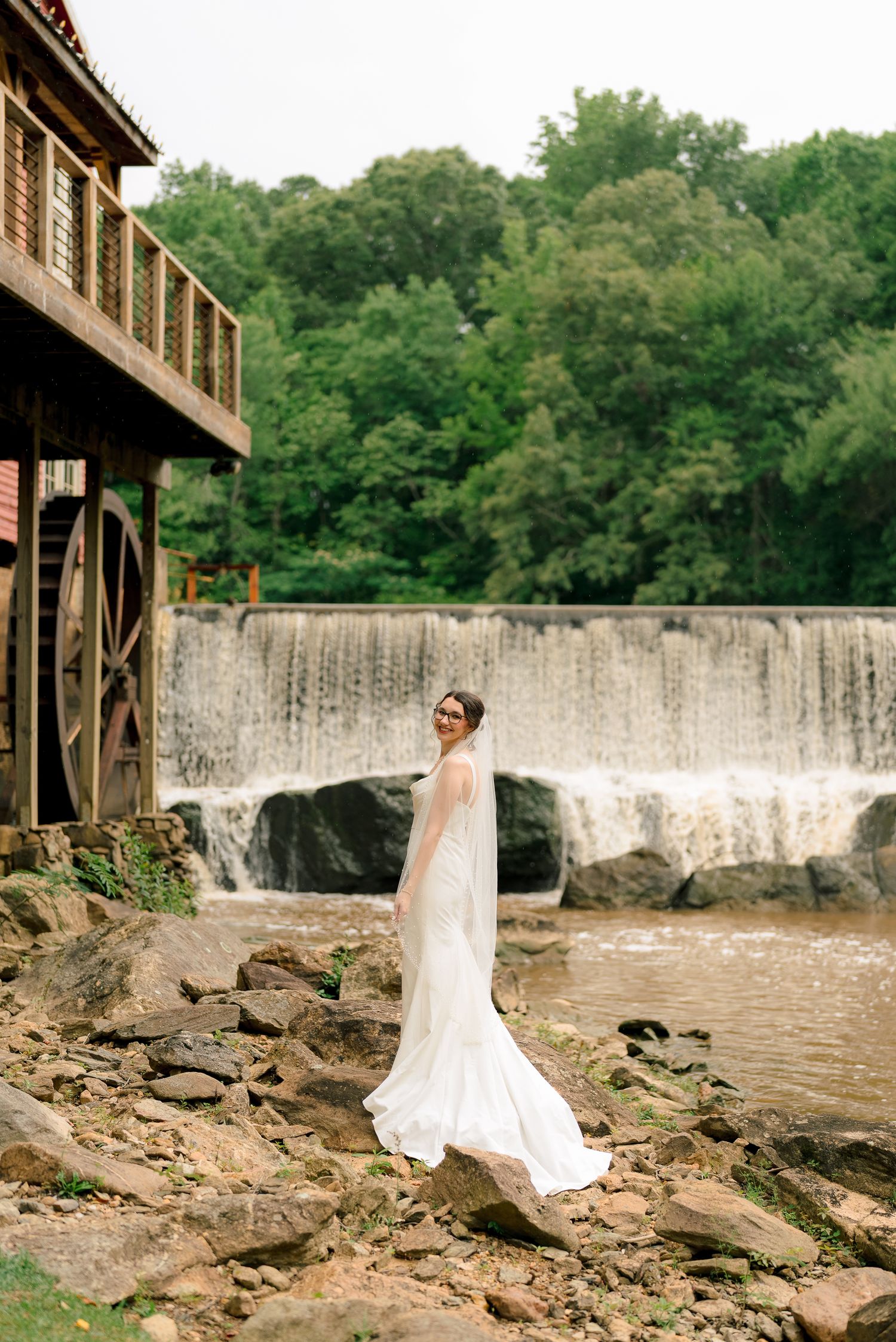 Elegant bride in white gown and veil standing on rocks beside waterfall and historic grist mill.