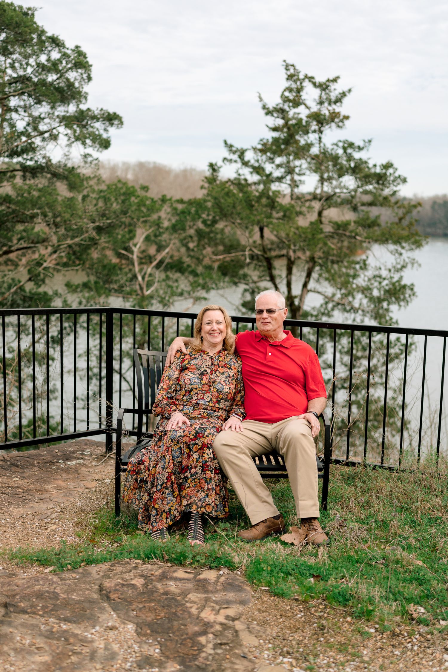 A couple sits on a bench overlooking a scenic river, surrounded by trees, the woman in a floral dress and man in a red polo.