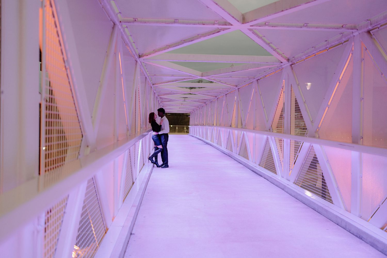 Couple sharing a romantic embrace on a purple-lit covered pedestrian bridge with geometric steel framework.
