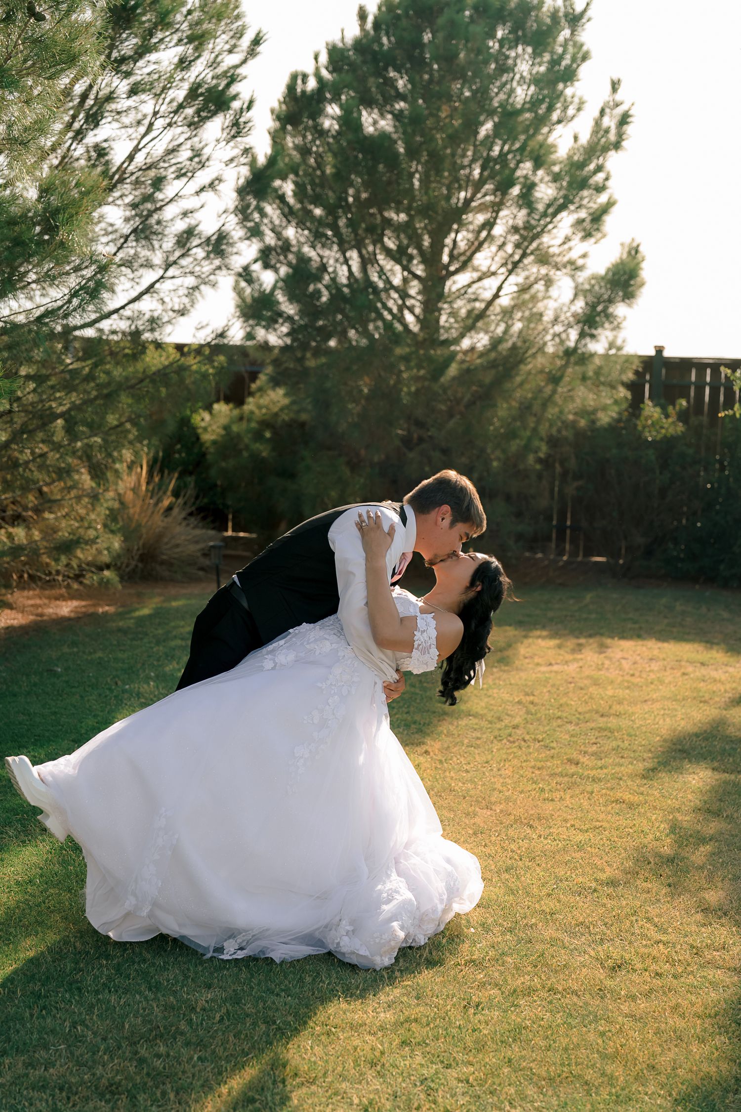 A groom dips his bride in a white ballgown for a romantic kiss on a green lawn surrounded by trees at golden hour.
