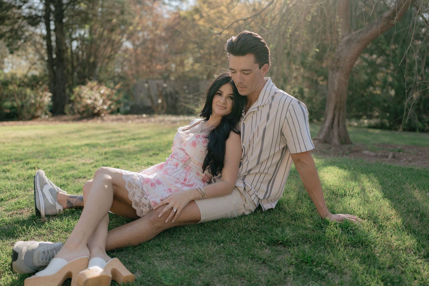 Couple sitting together on green grass in a sunny park, woman in floral dress leaning against man in striped shirt.
