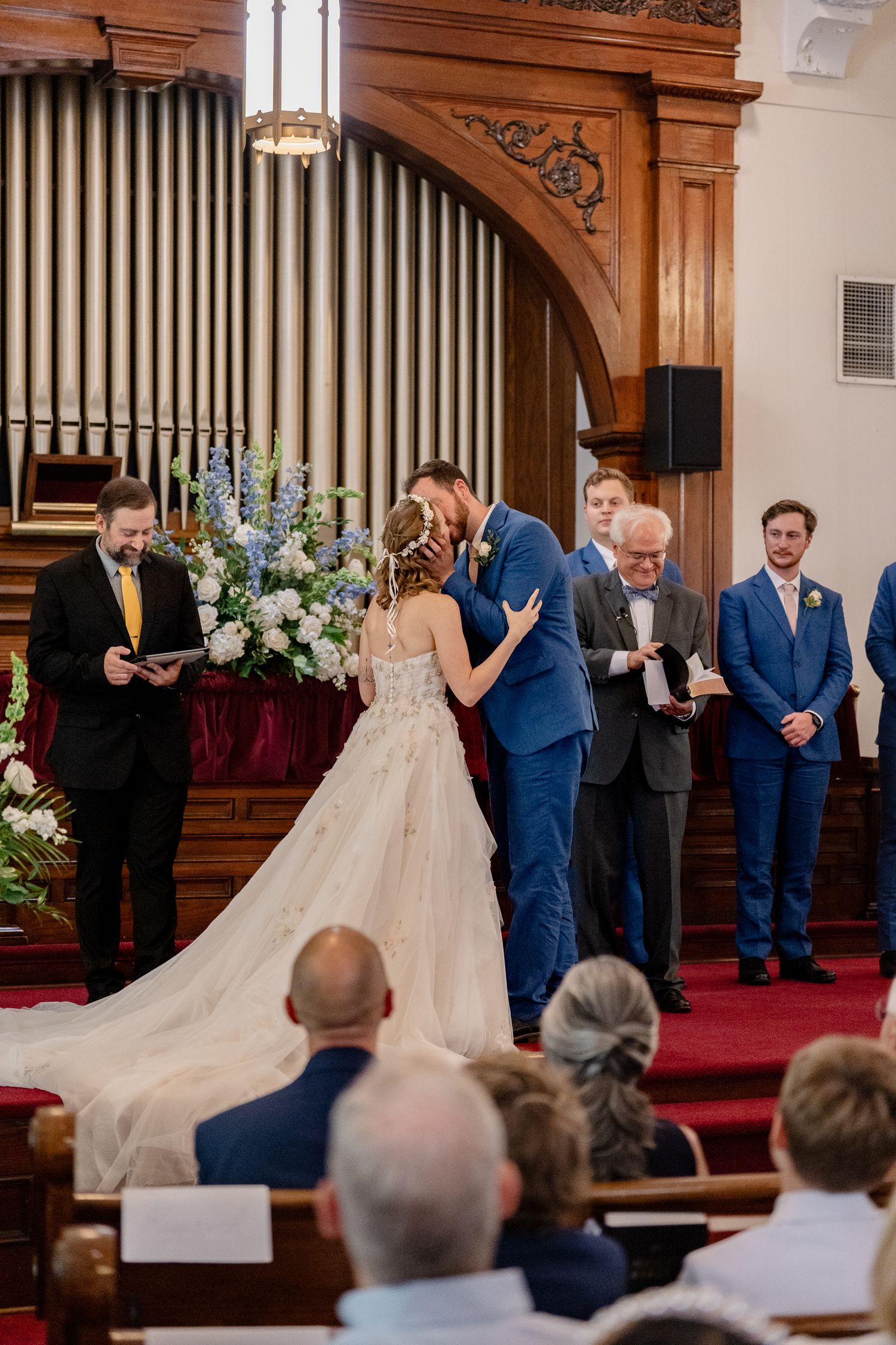 A wedding ceremony takes place at the altar of a traditional church with ornate wooden organ pipes in the background.