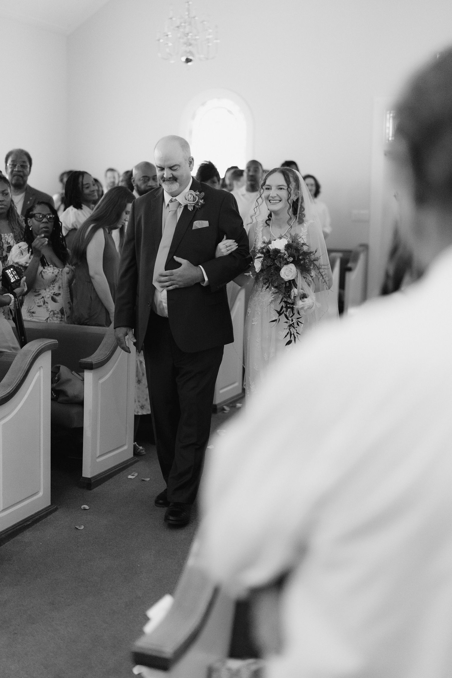 Black and white photo of smiling bride walking down church aisle on fathers arm holding bridal bouquet.