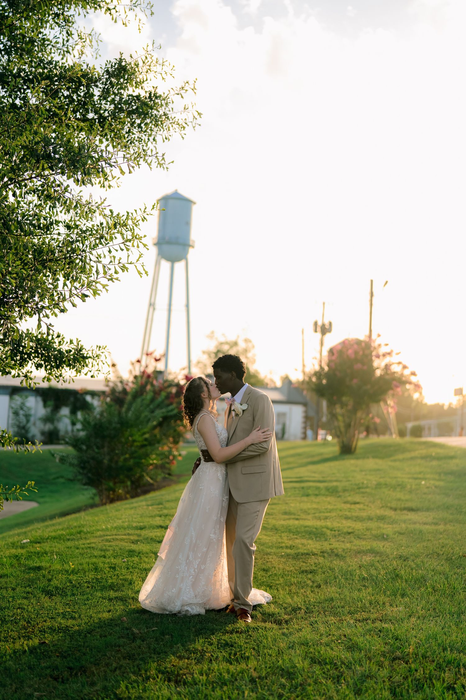 Bride and groom share a romantic embrace on green lawn at golden hour with water tower backdrop.