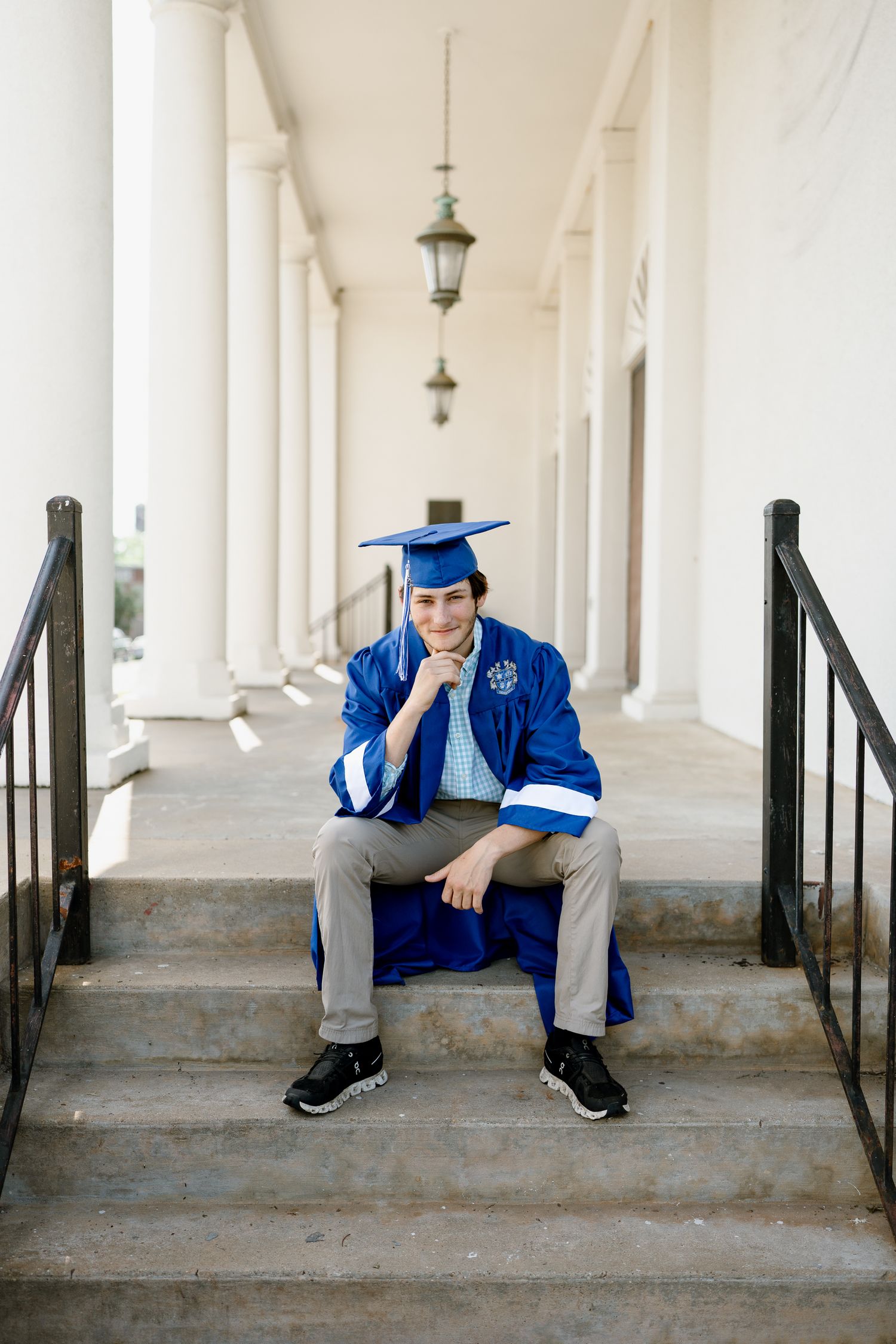Graduate in blue cap and gown sits contemplatively on stone steps between white columns.