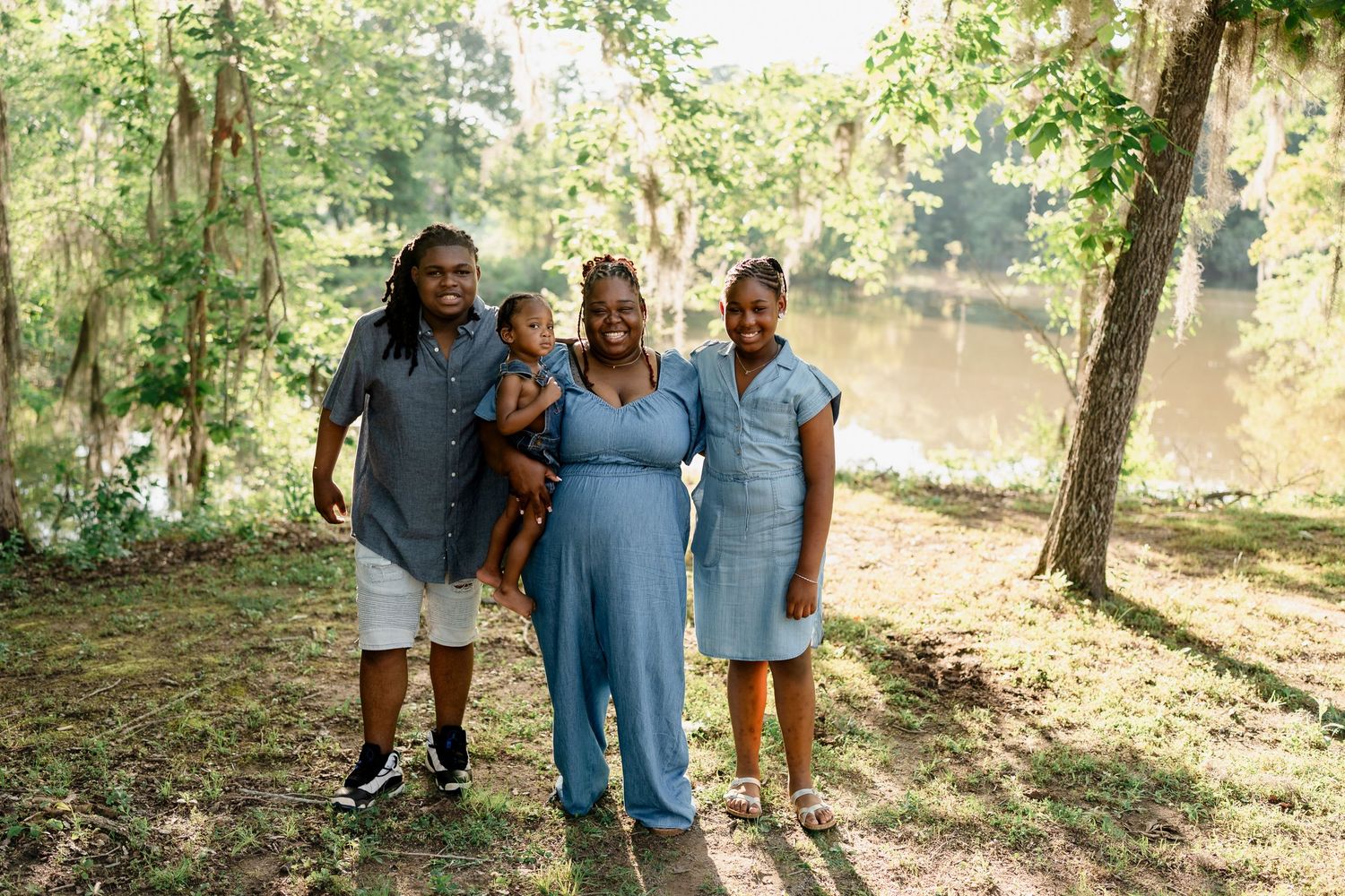 Family wearing coordinated blue outfits poses together in a sunny wooded area near water.
