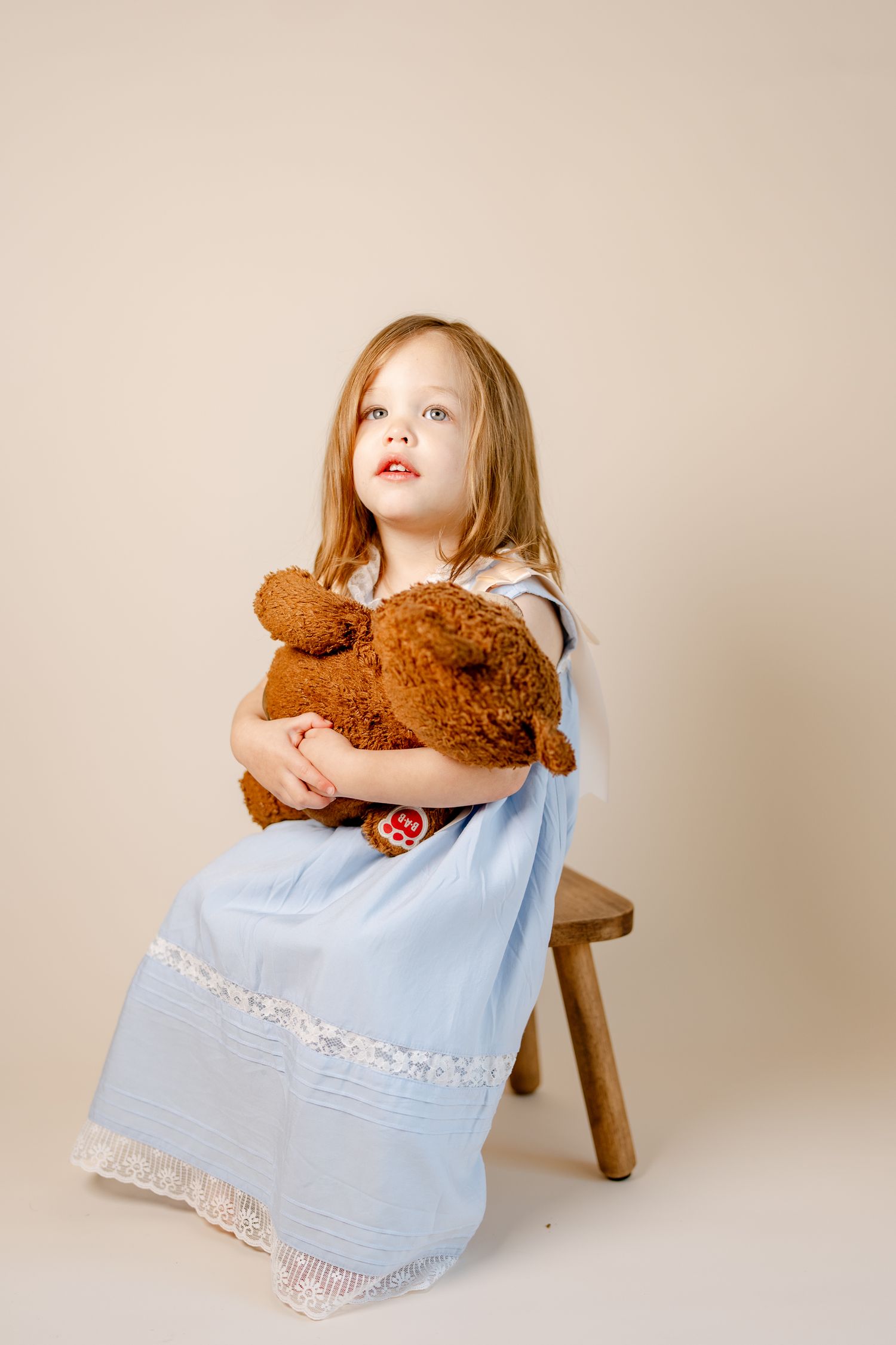 A light blue dress hugs a teddy bear against a neutral studio backdrop.