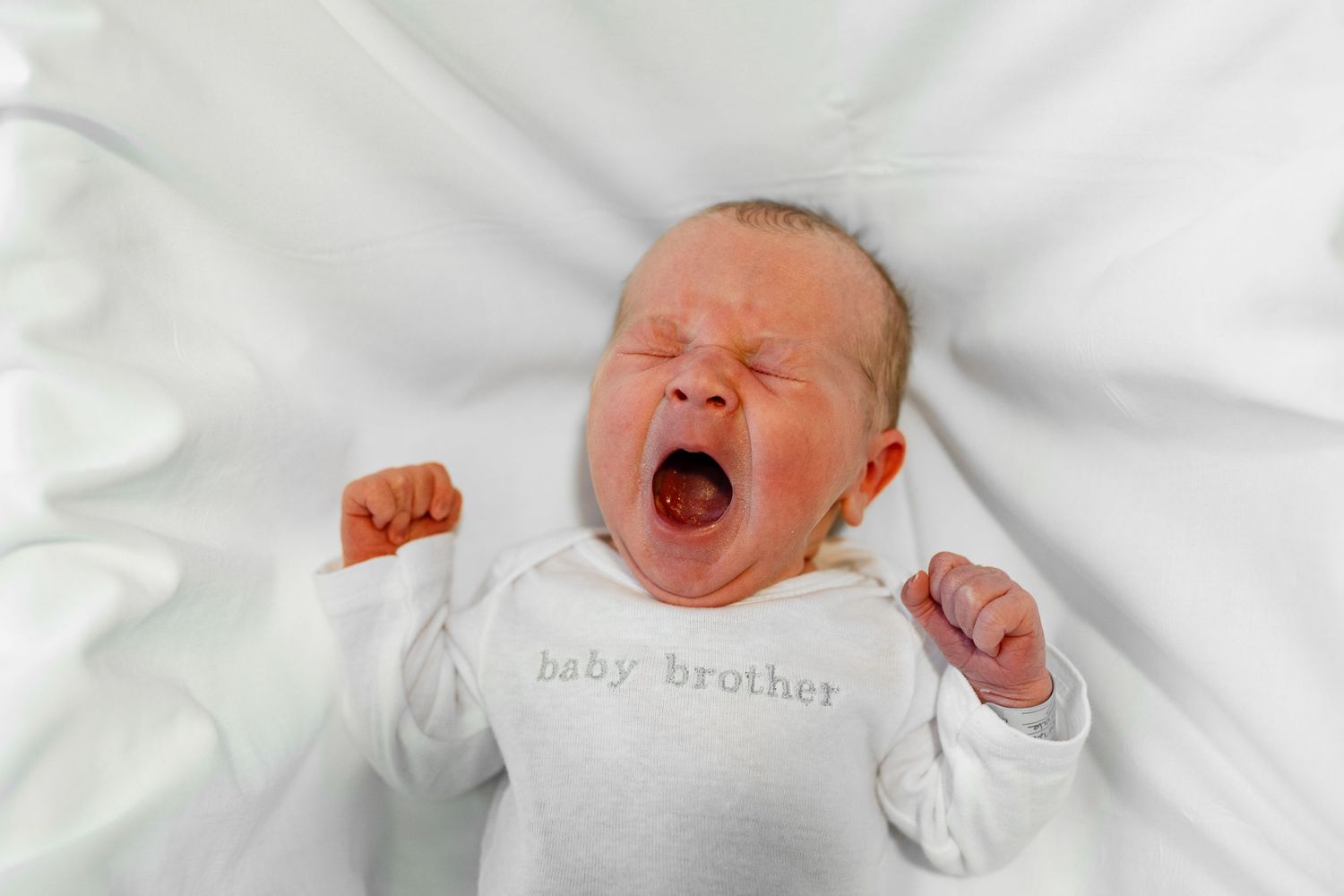 Infant wearing white clothing yawns while laying on crisp white bedding.