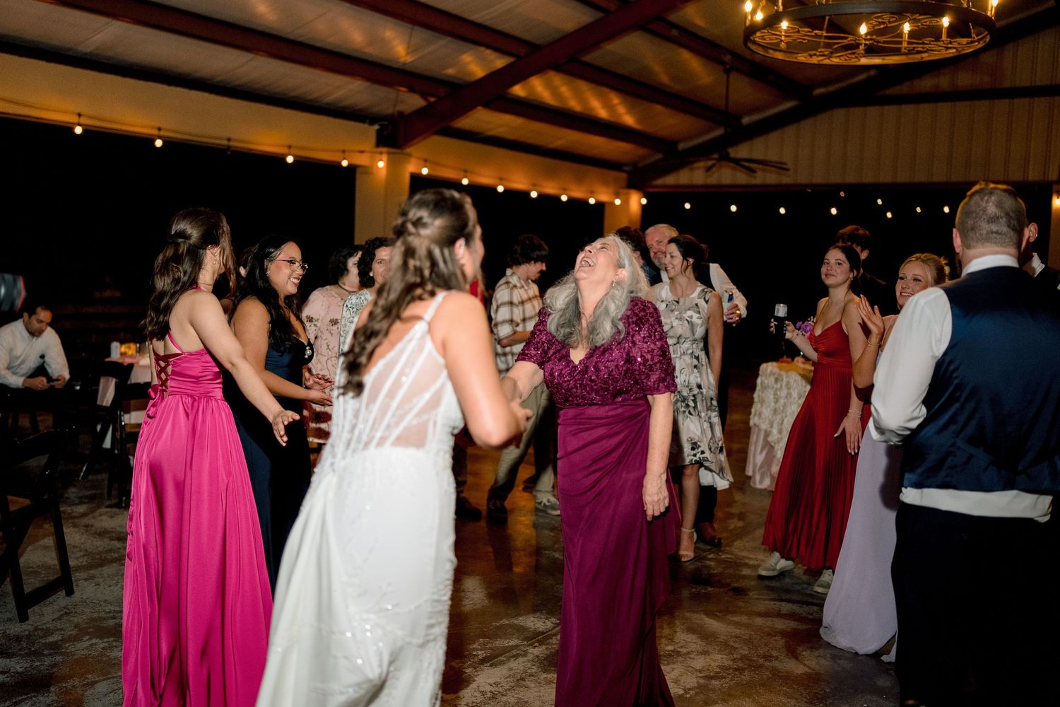 Wedding guests in colorful gowns dance with bride at lively outdoor evening reception under pavilion.