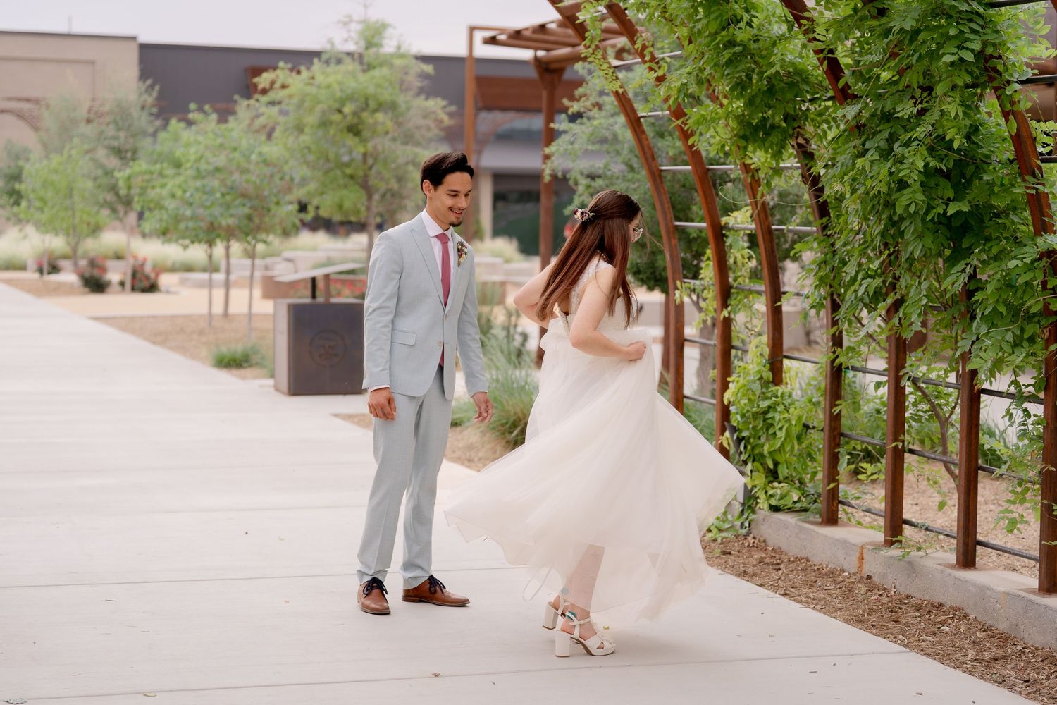 A couple in wedding attire walks along a winding path surrounded by desert landscaping and trees.