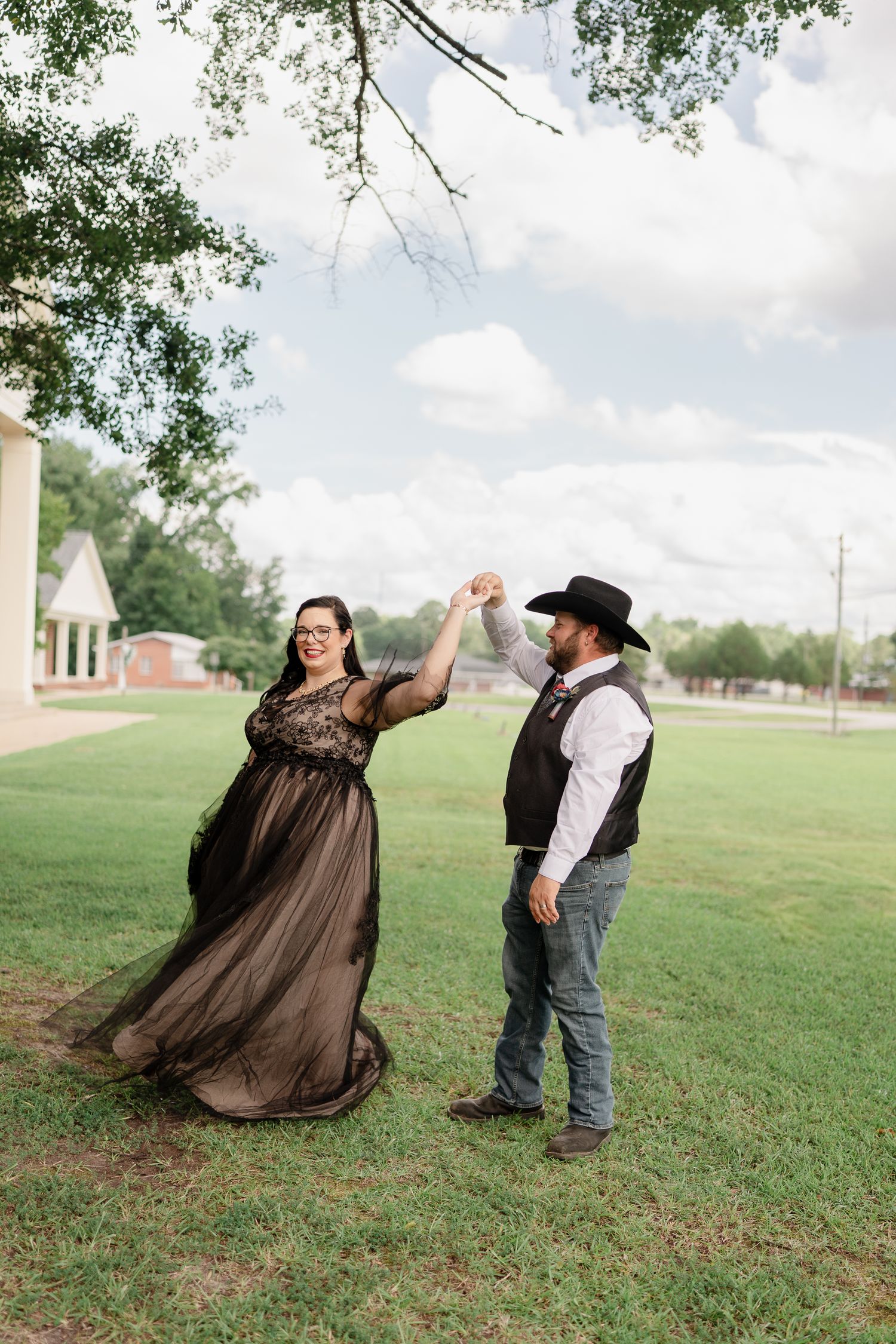 A couple in western attire dances playfully on a grassy field with trees in the background.