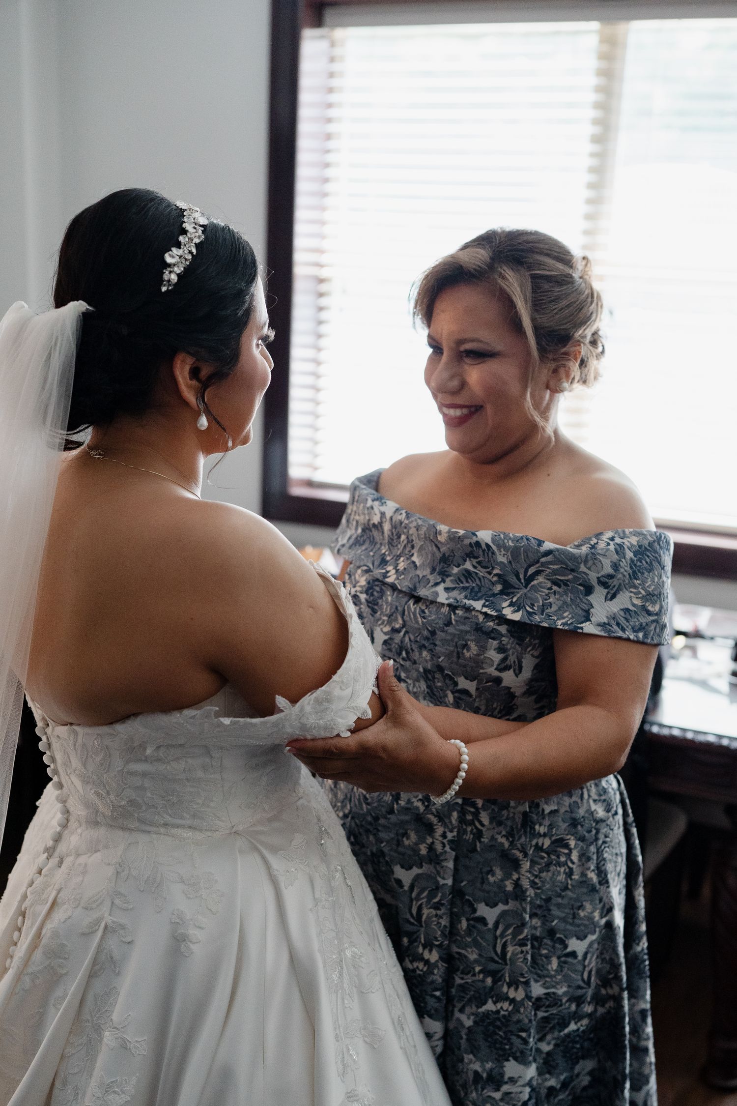 Two women share an emotional moment before a wedding, with one wearing a white gown and the other in a blue floral dress.