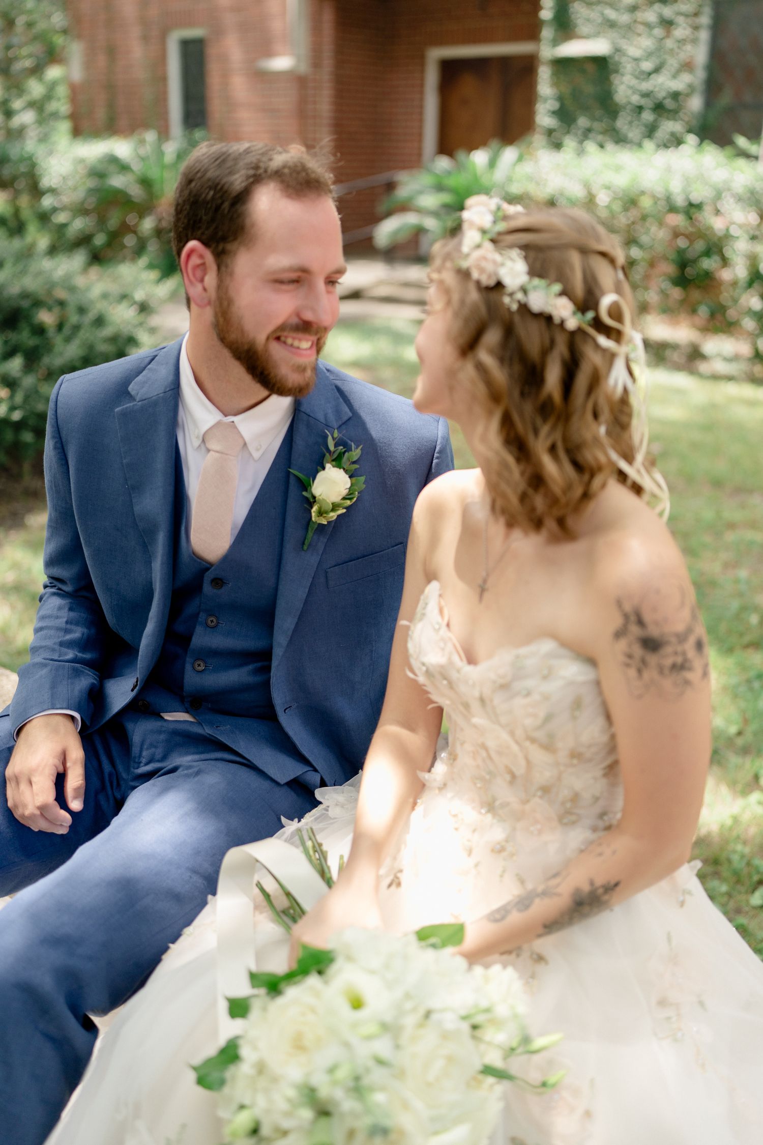 A couple in wedding attire shares playful moments together in a garden setting with brick buildings behind them.