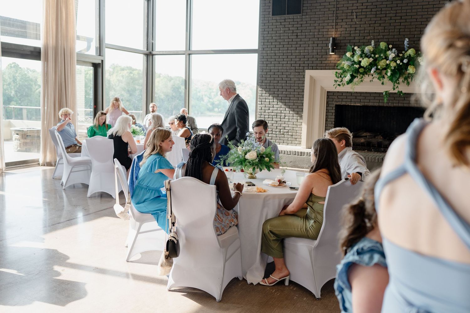 Wedding guests mingle in an elegant reception venue with floor-to-ceiling windows and white chair covers.