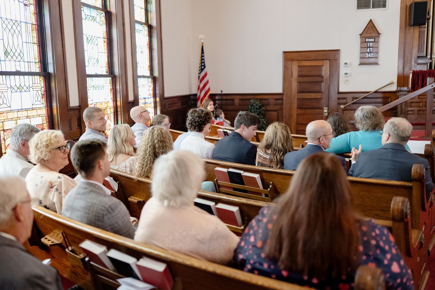 Church congregation seated in wooden pews with stained glass windows visible in the background.