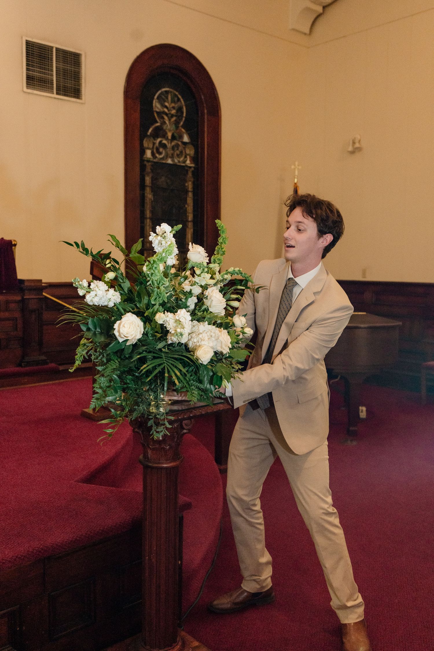 A person in a light suit arranges white floral decorations on wooden church pews.