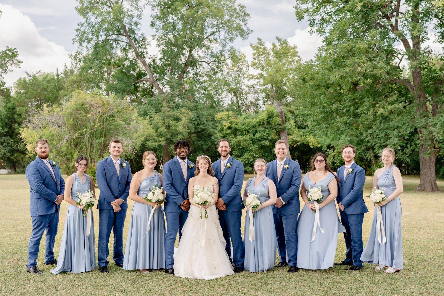 Wedding party lounges on grassy hill wearing coordinated light blue and navy attire in outdoor setting.