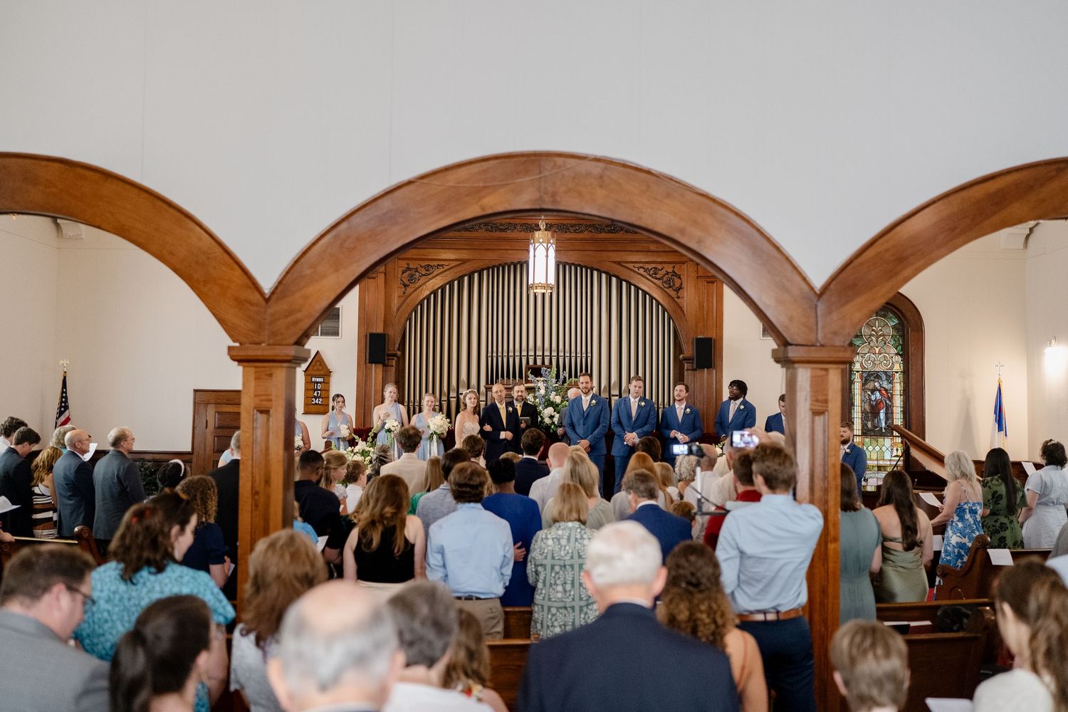 Wide angle view of wedding ceremony attendees gathered in ornate church sanctuary with wooden arches.