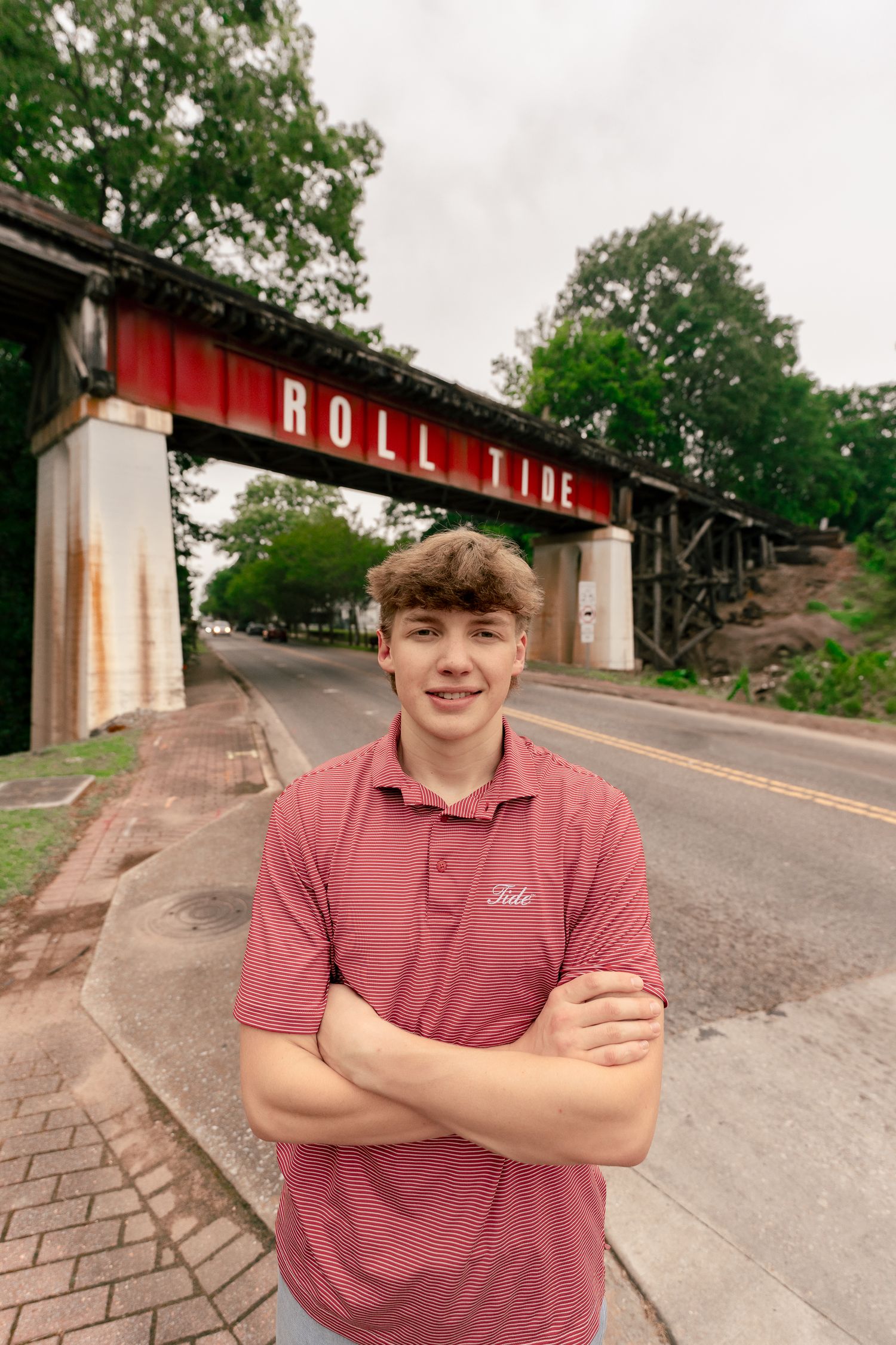 A person stands near a bridge with 'Roll Tide' signage and green trees in the background.