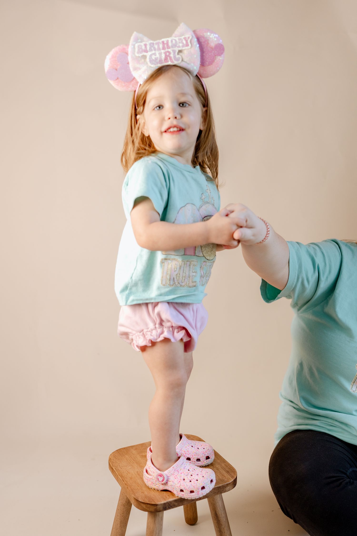 A small wooden stool and aqua t-shirt are featured against a beige backdrop with pink accessories.