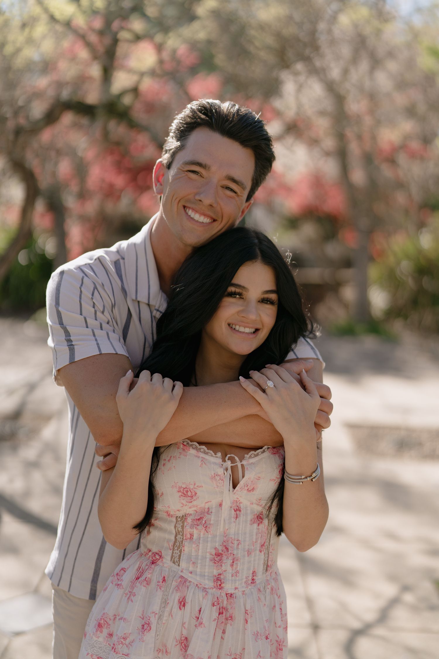 A smiling couple embraces outdoors, man hugging woman in floral dress, with blooming pink trees in background.