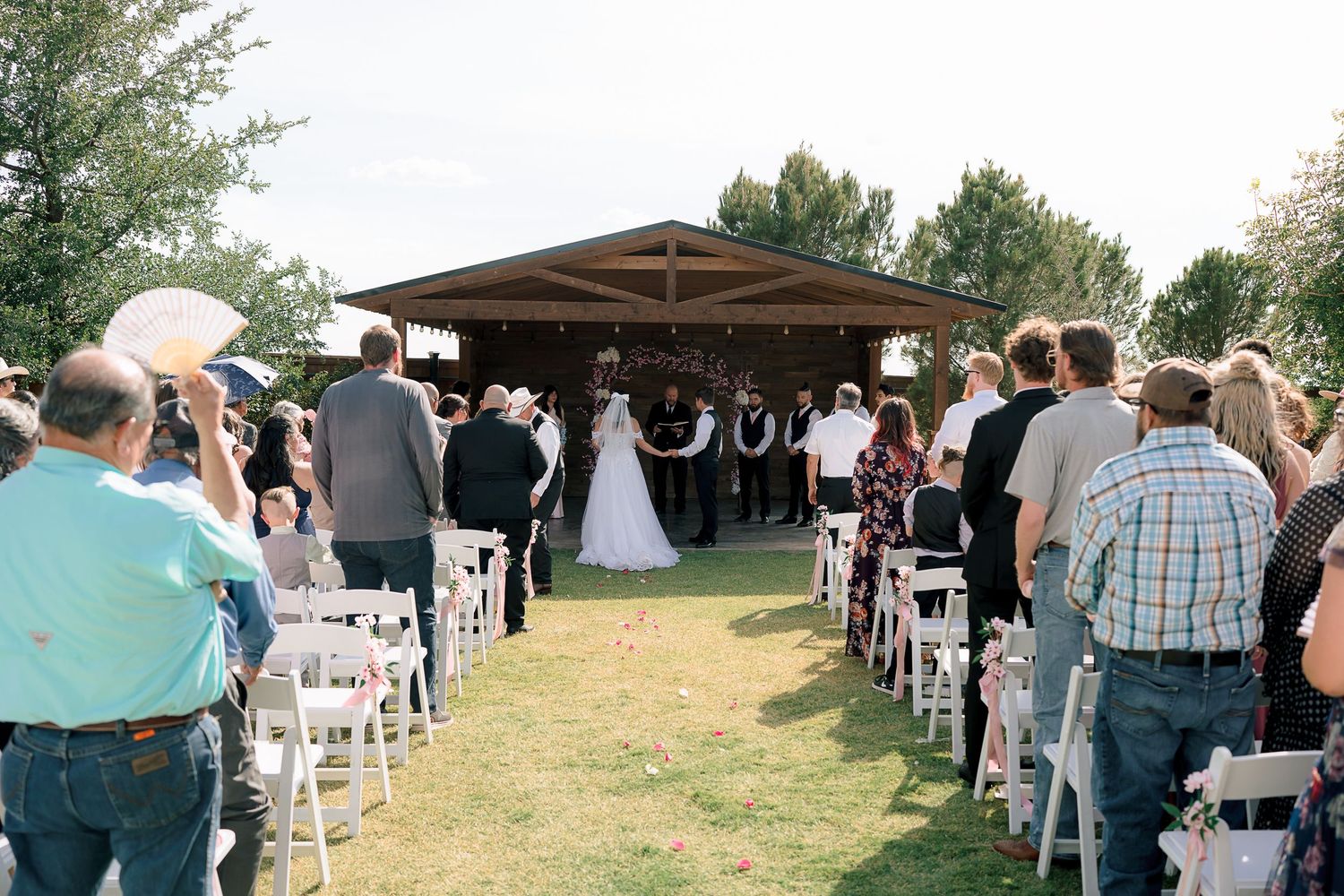 Outdoor wedding ceremony with guests seated in white chairs facing bride and groom under a wooden pavilion.